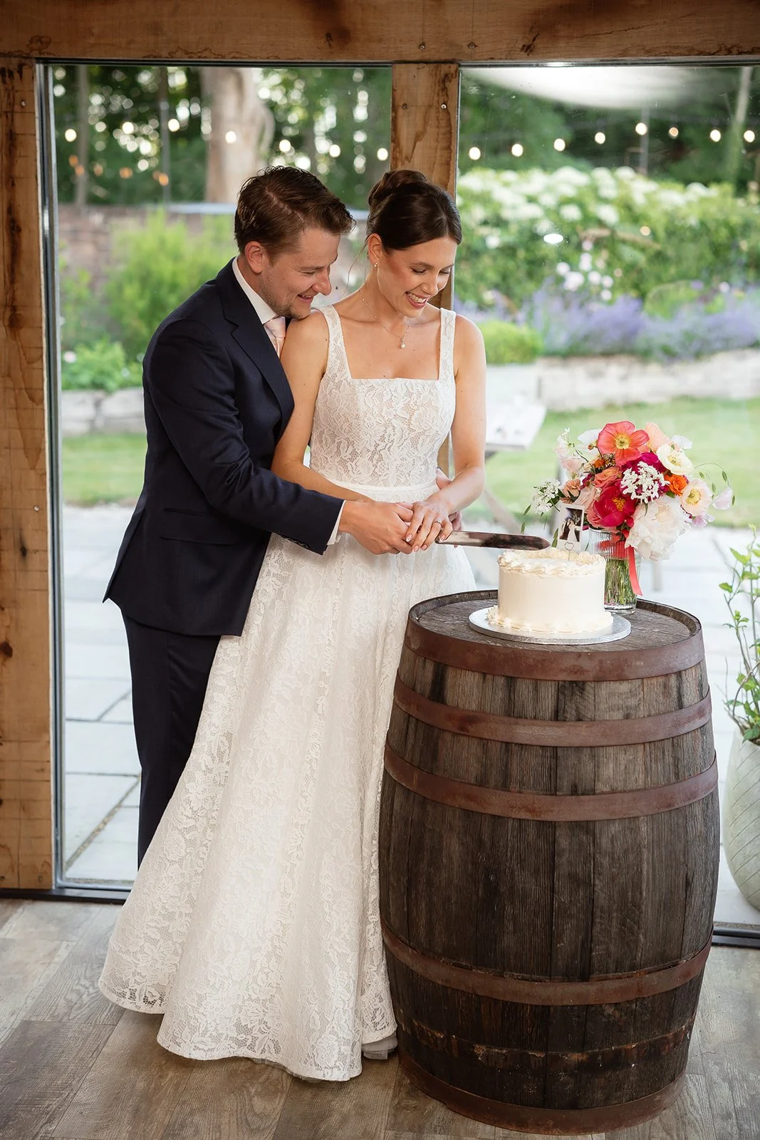 A bride and groom in wedding attire sharing a cake cutting ceremony during their wedding reception outdoors, with a flower arrangement on a wine barrel table beside them.