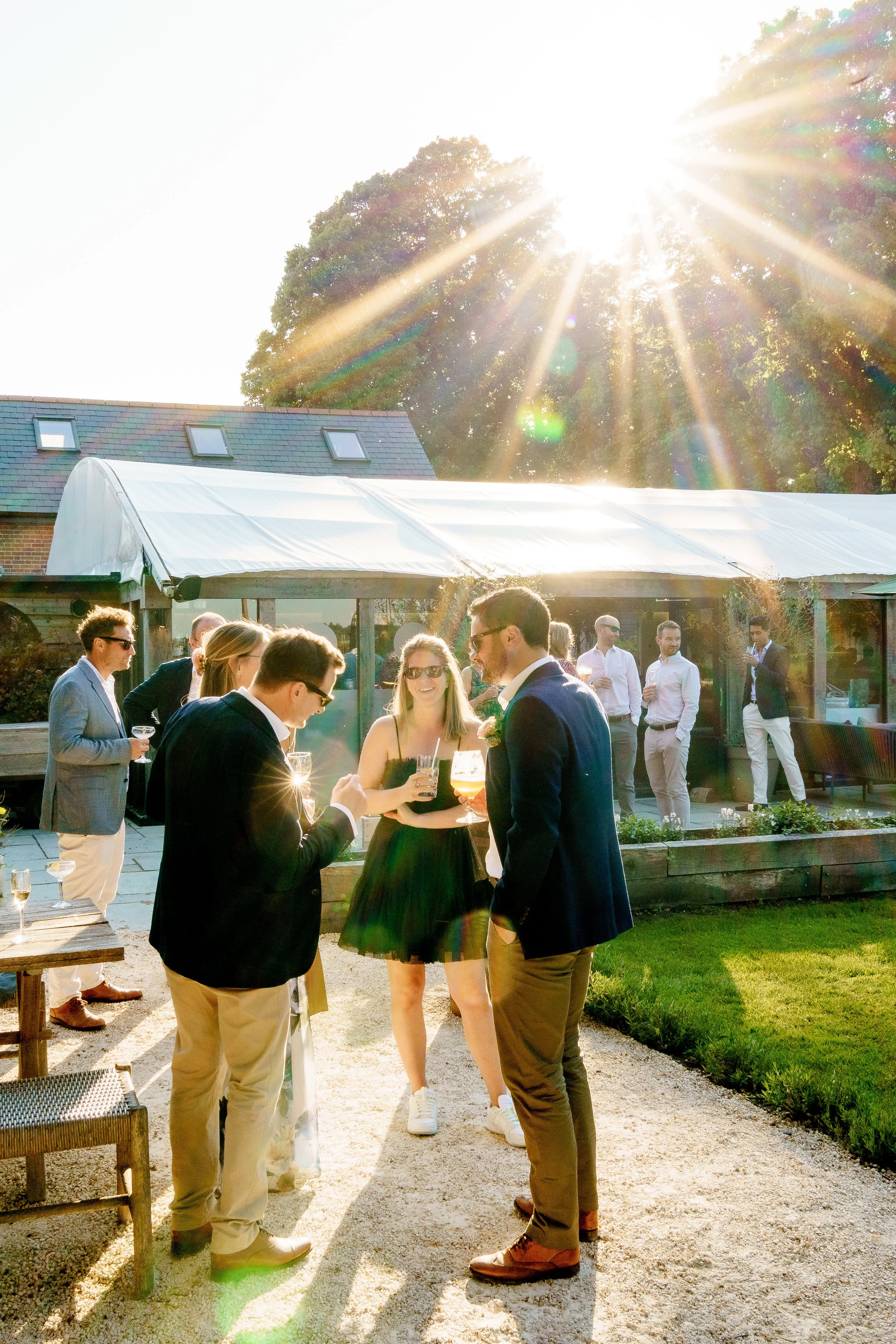 People socializing outdoors in the late afternoon sunlight, holding drinks, in a garden with a greenhouse or outdoor structure.