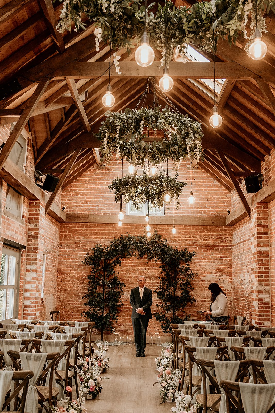 Rustic indoor wedding venue with brick walls, wooden chairs, floral aisle decor, hanging greenery, and exposed bulb lighting.