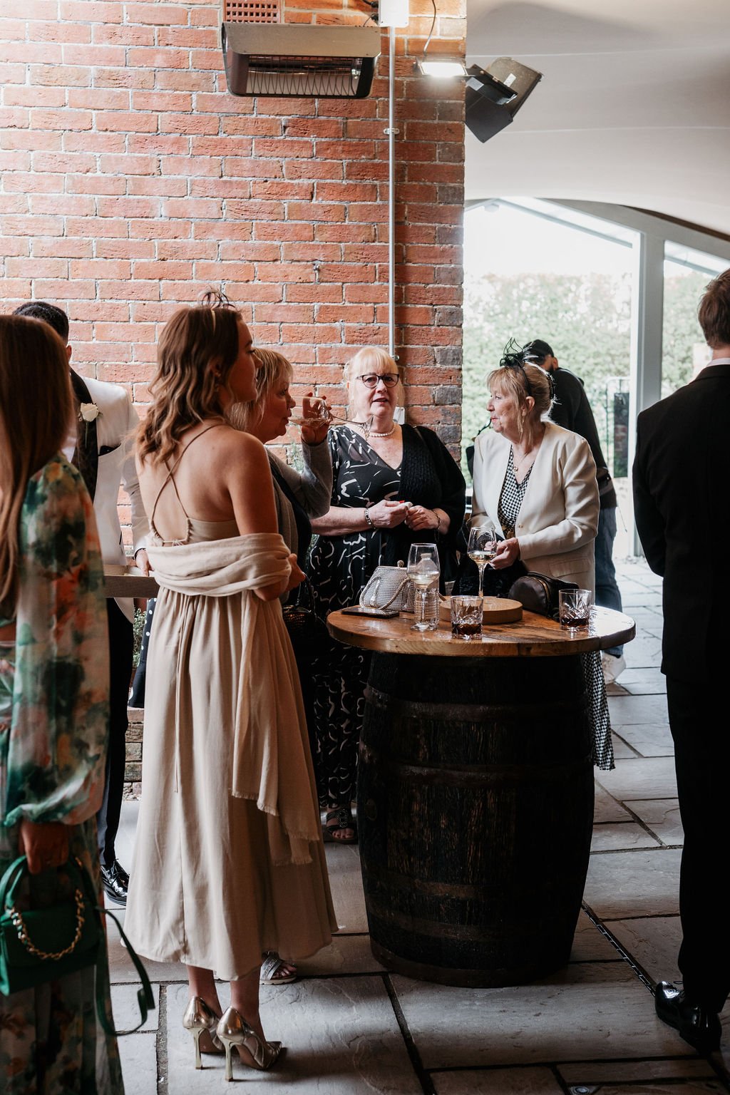 Group of people socializing at an indoor event, standing around a wooden barrel table with drinks, near a brick wall.