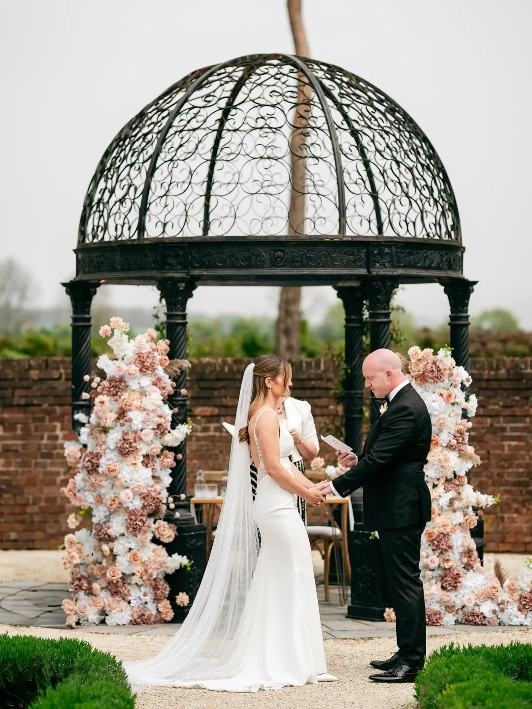 A bride and groom exchange vows during a wedding ceremony outdoors under a decorative metal gazebo with floral arrangements on the sides.