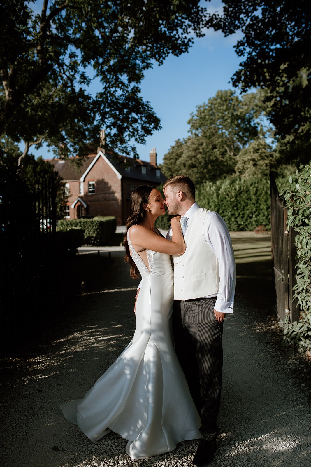 A bride and groom stand close together, touching foreheads and smiling, outdoors with trees and a brick house in the background. The bride wears a white wedding gown and the groom a white shirt with a vest and dark pants. Bright sunlight creates shad