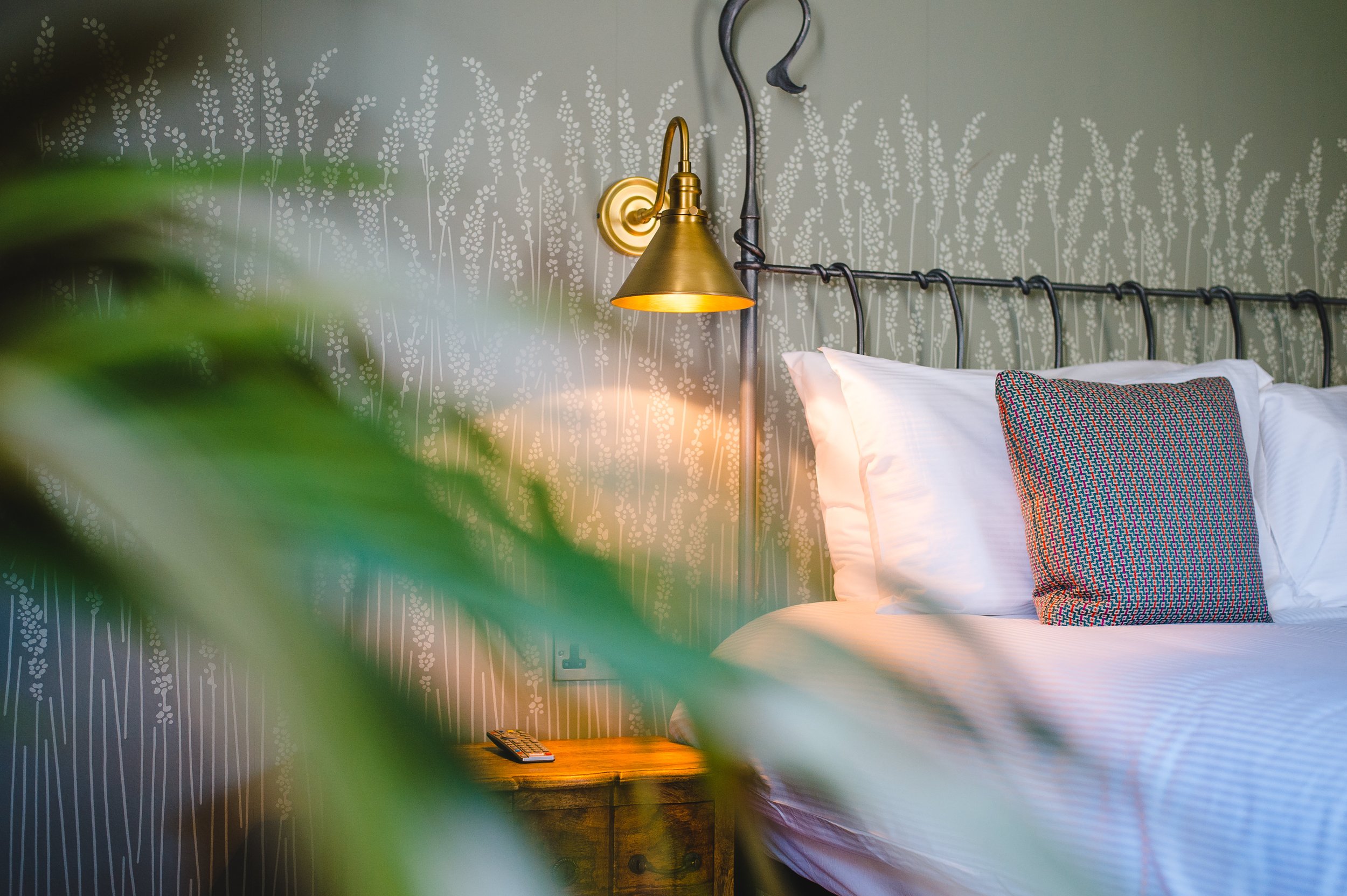 Cozy bedroom interior with a metal bed frame, white bedding, and a patterned pillow. A brass wall sconce is lit above a wooden nightstand, partially obscured by green plant leaves.