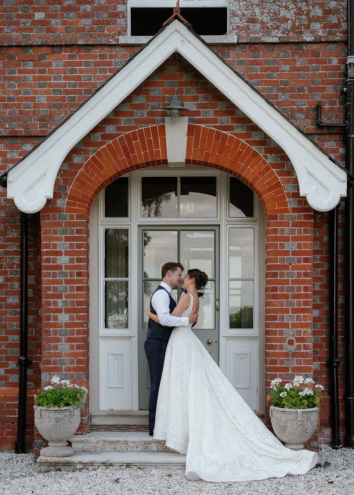 A newlywed couple standing close and about to kiss in front of a vintage brick house with white trim and a large window, surrounded by potted flowers.