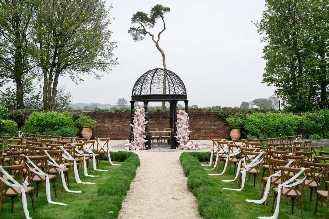 An outdoor wedding setup with an aisle leading to a decorative gazebo, adorned with pink and white flowers, in a lush green garden.