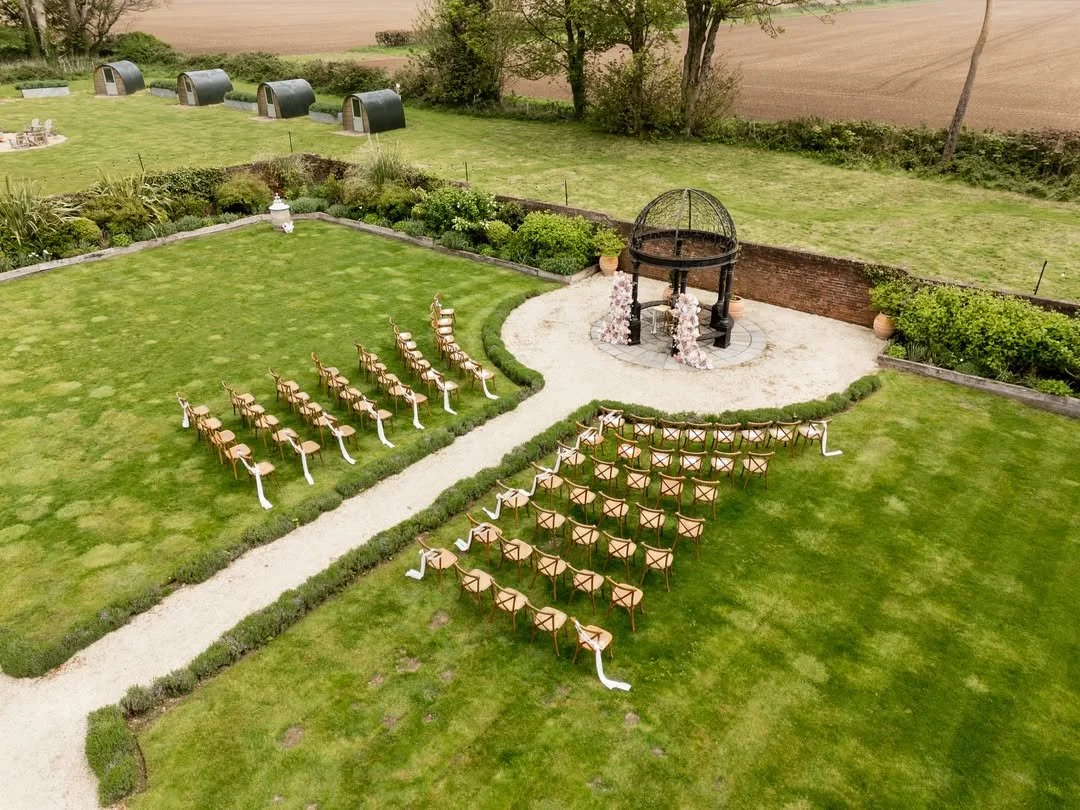 Outdoor wedding ceremony setup with rows of wooden chairs, a gazebo, and floral decorations on a grassy lawn.