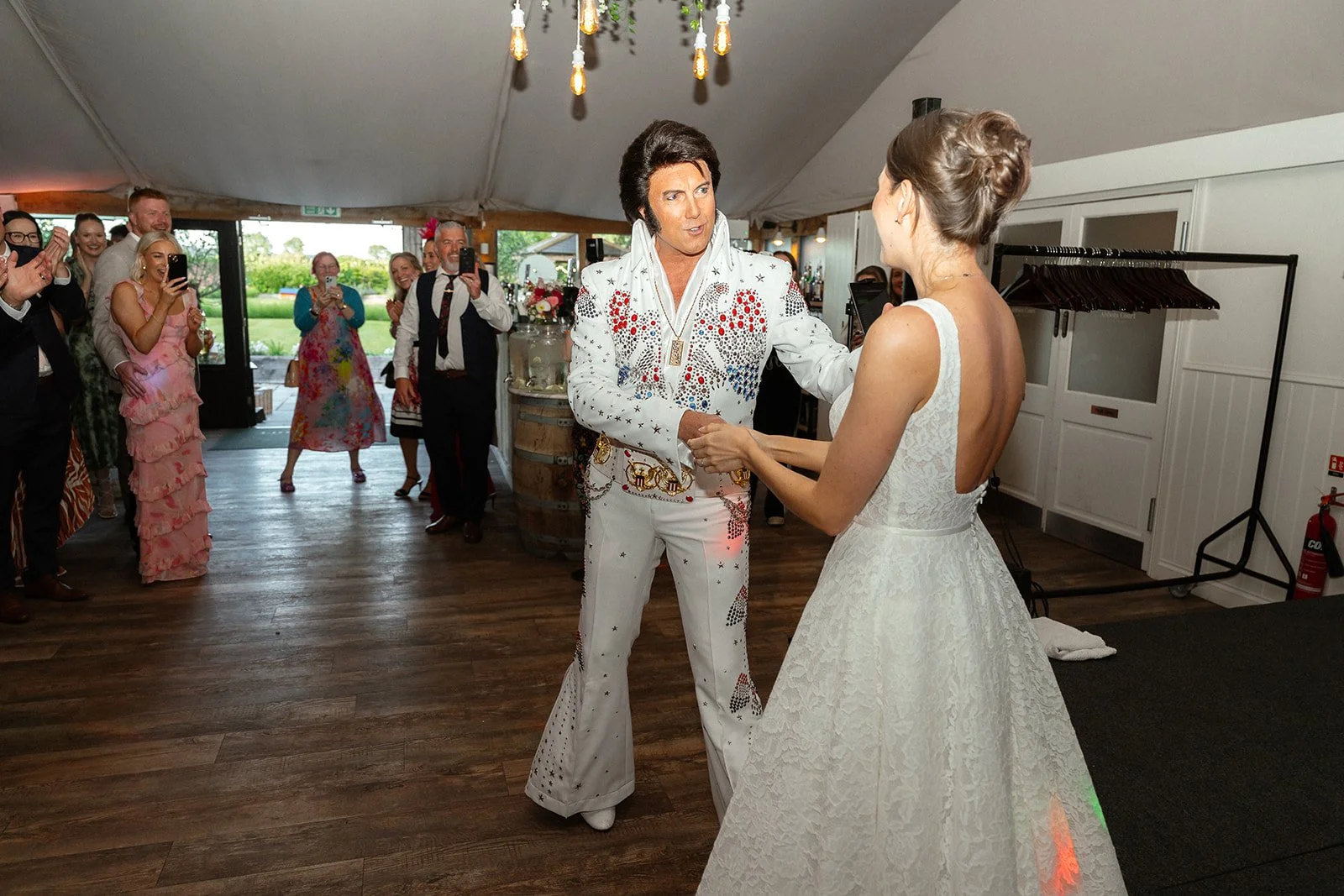 A person dressed as Elvis Presley at a wedding dance, holding hands with a bride, while guests watch and take photos.