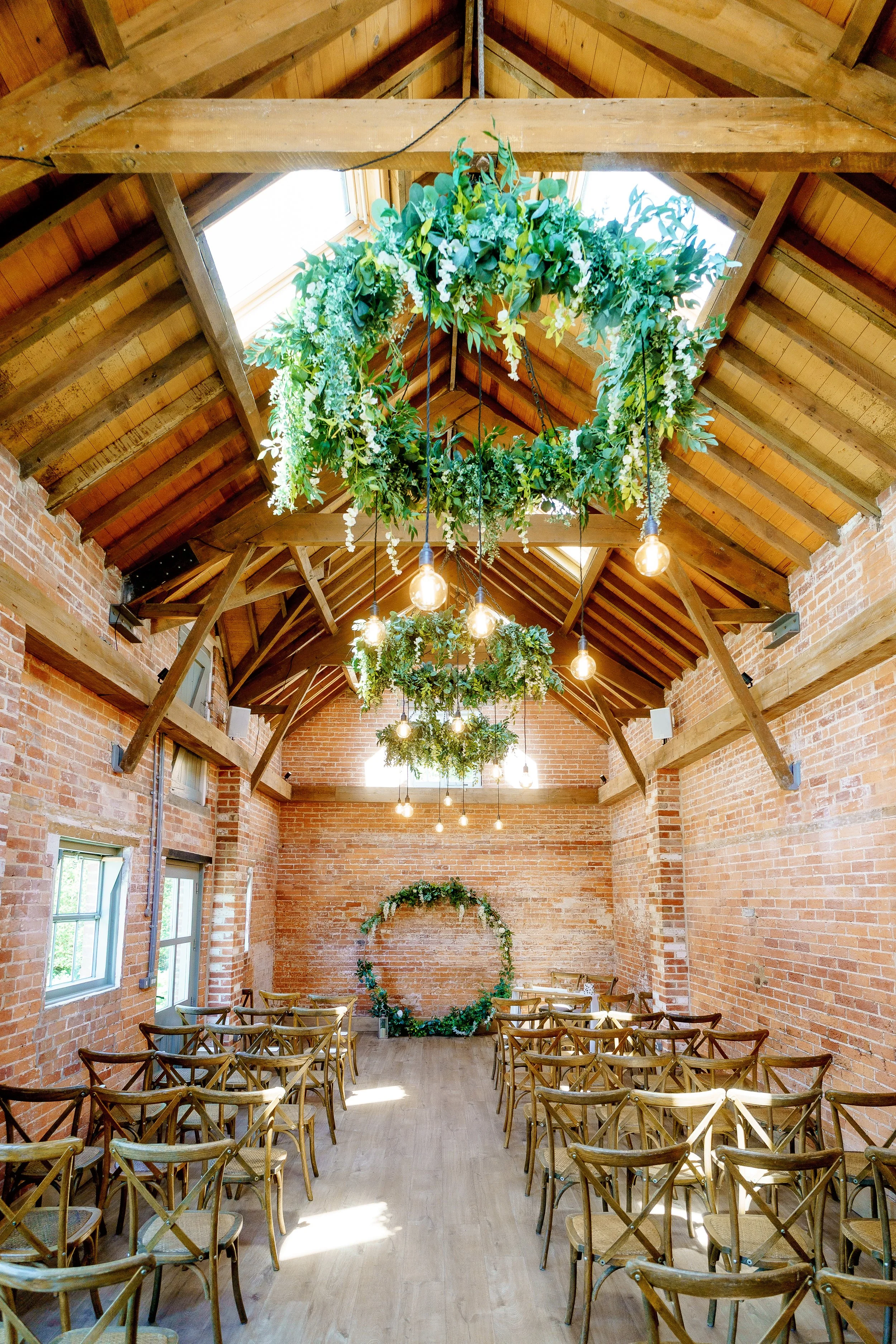 Indoor wedding venue with brick walls, wooden ceiling, and natural light. Decor includes hanging green foliage chandeliers and a large leafy wreath at the altar.