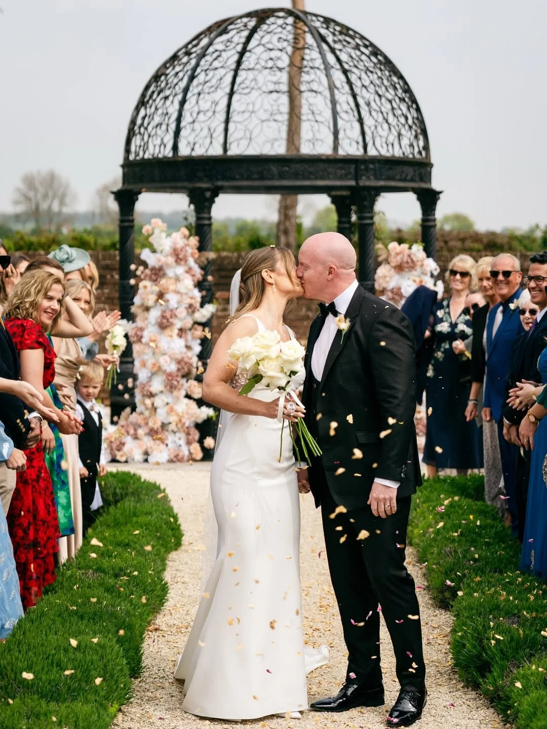 A bride and groom kiss during their outdoor wedding ceremony, surrounded by friends and family, with floral decorations and a black ornate gazebo in the background.