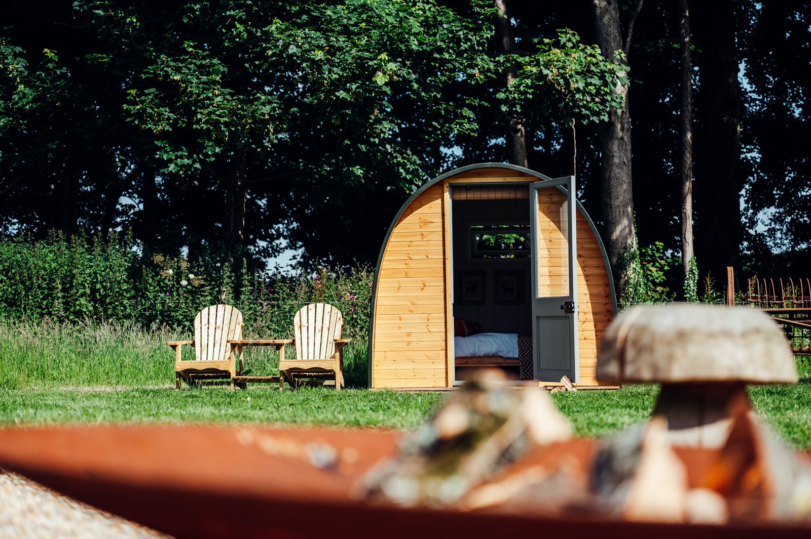 A wooden glamping pod with an open door, two Adirondack chairs outside, surrounded by grass and trees.