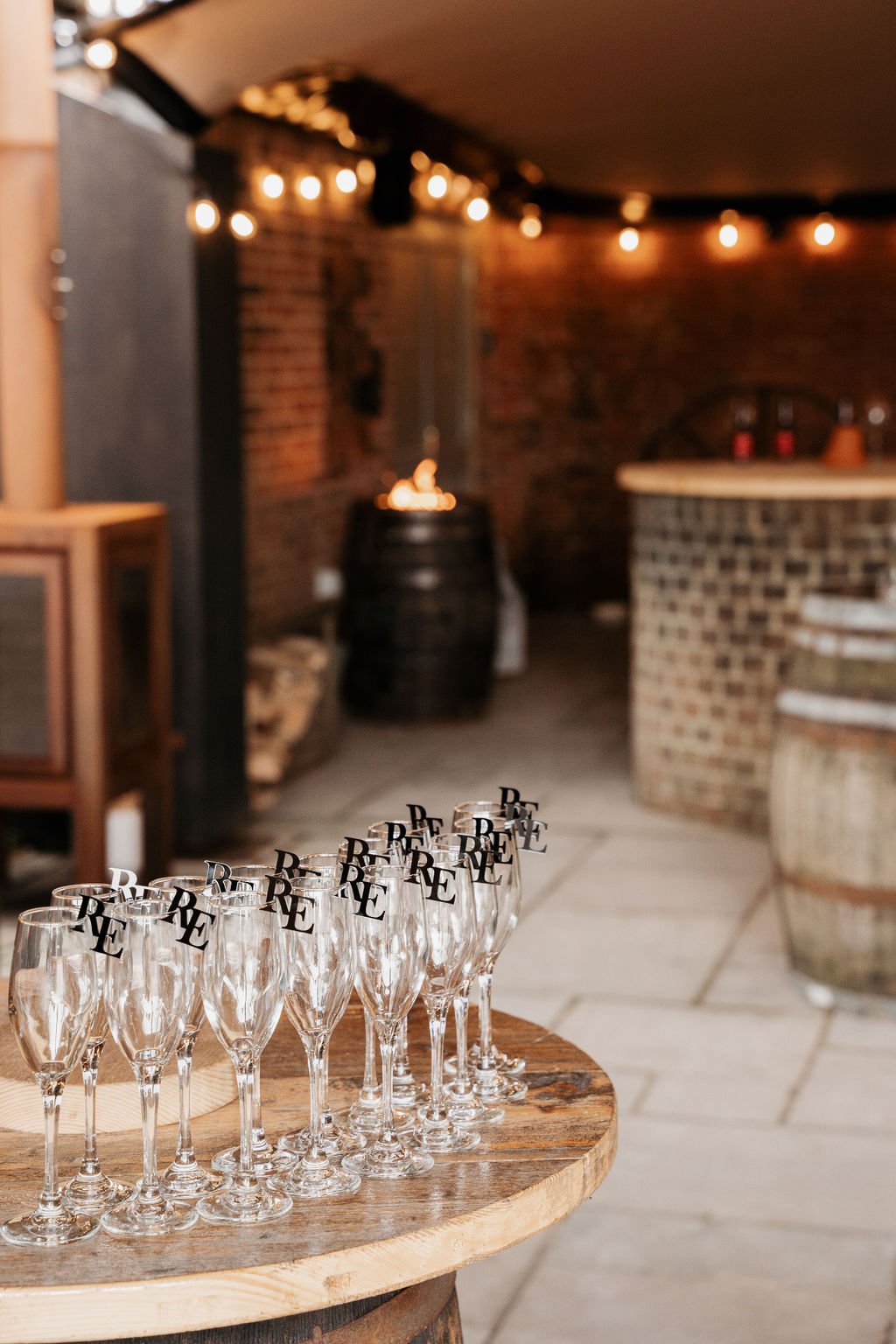 Rustic bar setting with empty champagne glasses on a wooden barrel table, string lights, and a brick wall in the background.