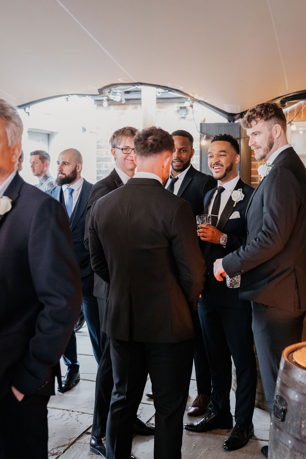 Group of men in suits at a social gathering, some holding drinks, engaged in conversation indoors.