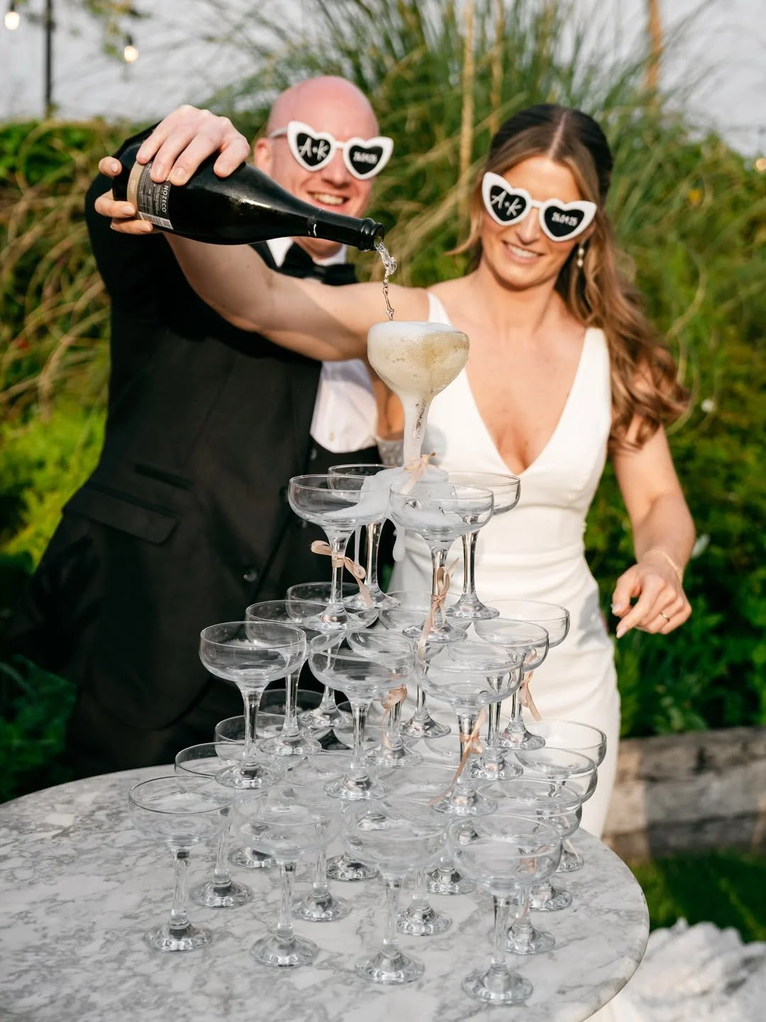 A couple at a celebration pouring champagne into a pyramid of glasses outdoors, both wearing heart-shaped sunglasses with message labels.
