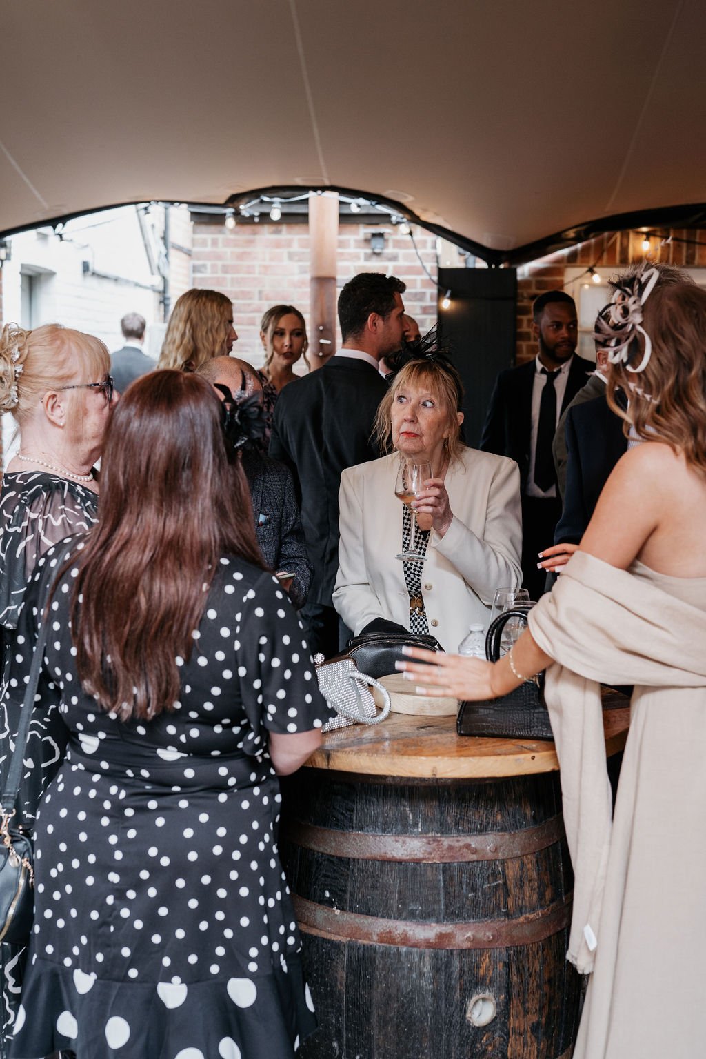 Group of people gathered around a barrel table at a social event, dressed in formal attire, with a woman holding a glass.