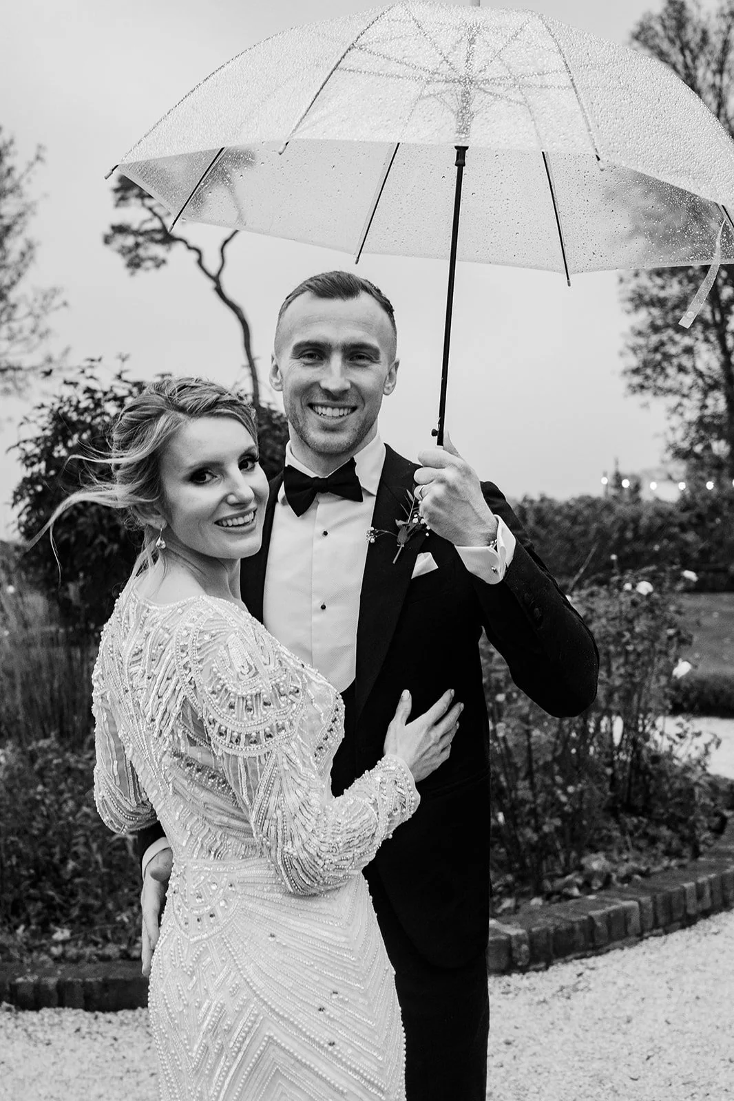 A black and white photo of a newlywed couple outdoors on a rainy day. The groom is holding a transparent umbrella over the bride. They are smiling and embracing, with a garden and trees in the background.