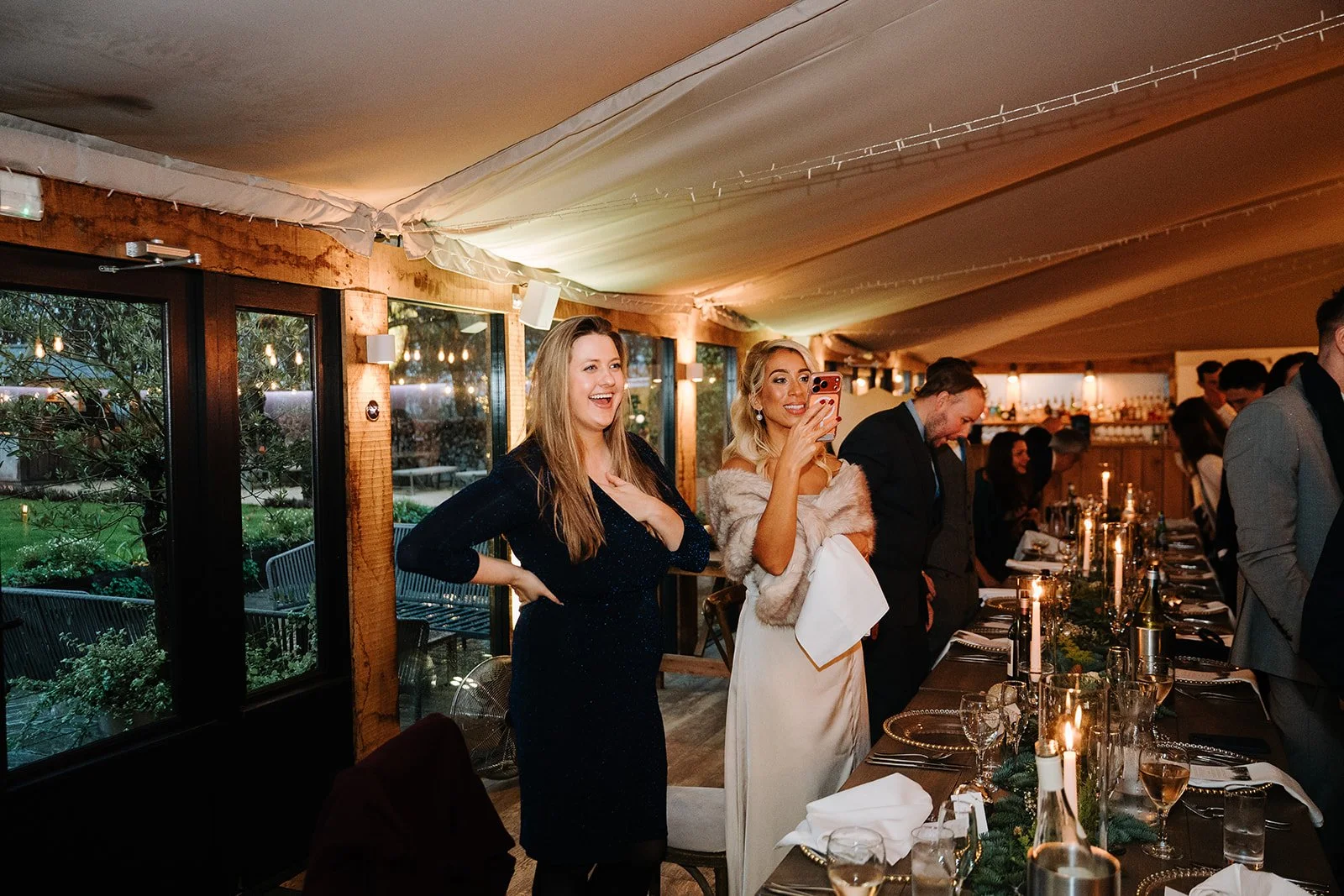 People at a formal dinner event, some standing and some bowing their heads, in a warmly lit room with a long table set for a celebration.
