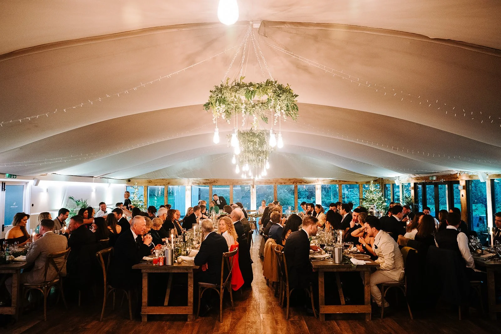 People seated at long tables in a decorated event tent, dining and socializing, with a chandelier and greenery hanging from the ceiling.
