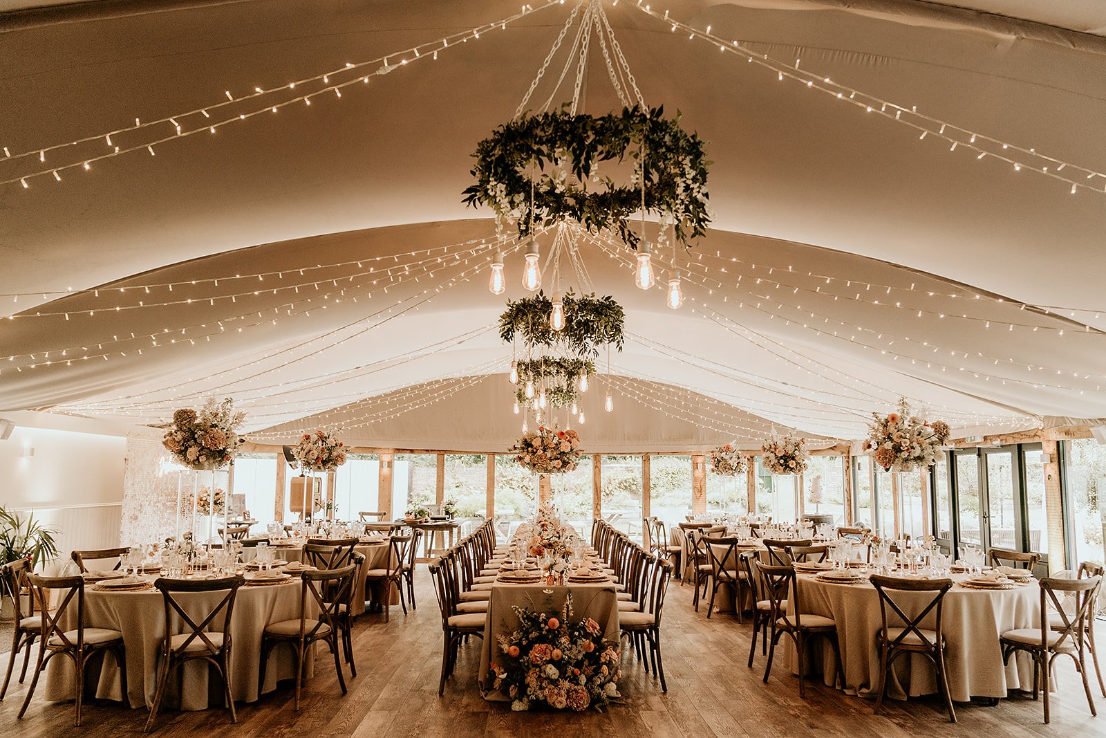 Elegant wedding reception setup in a marquee tent with string lights, floral centerpieces, and round tables.