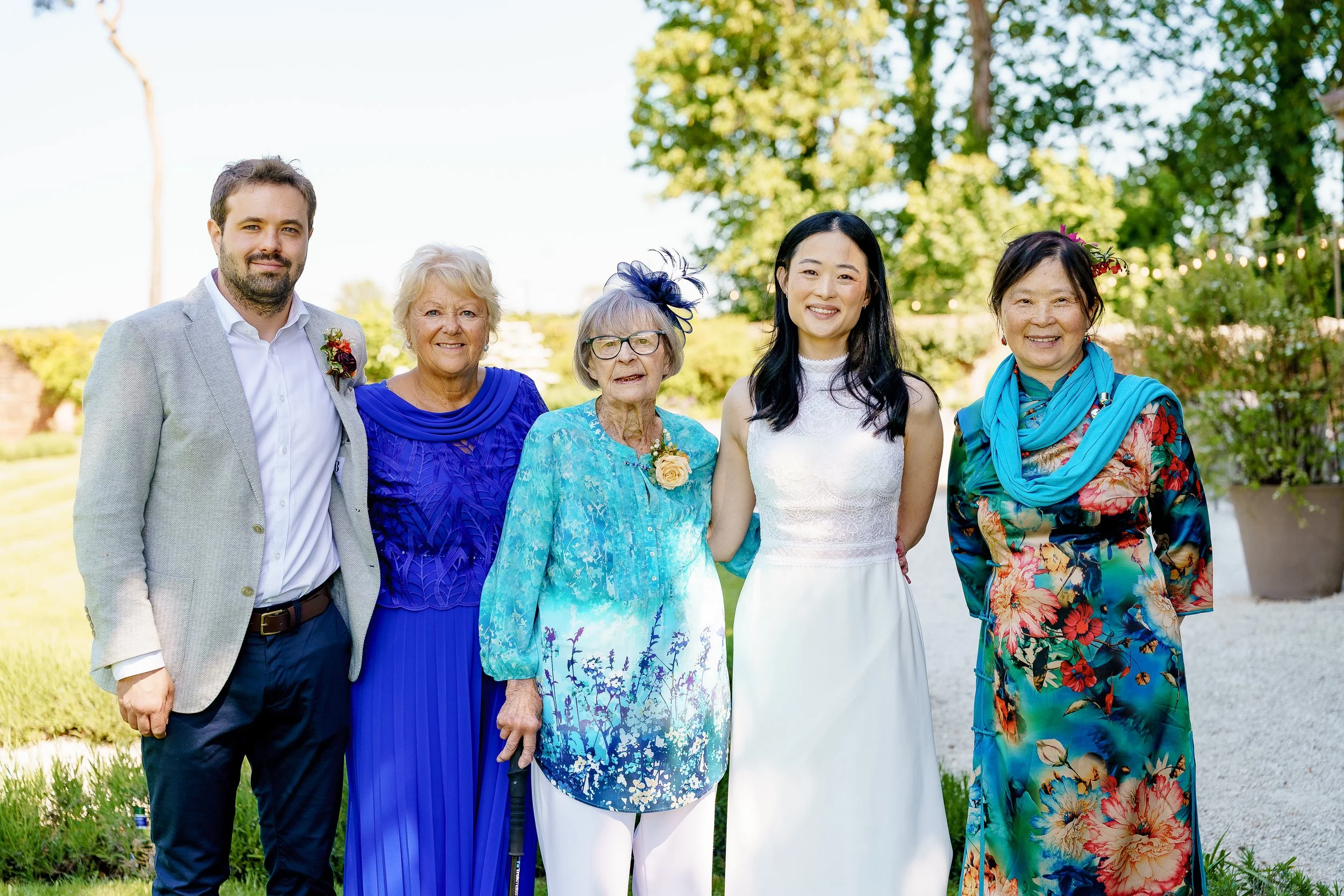 Group of six people standing outdoors during a wedding celebration, with trees and greenery in the background, smiling at the camera.