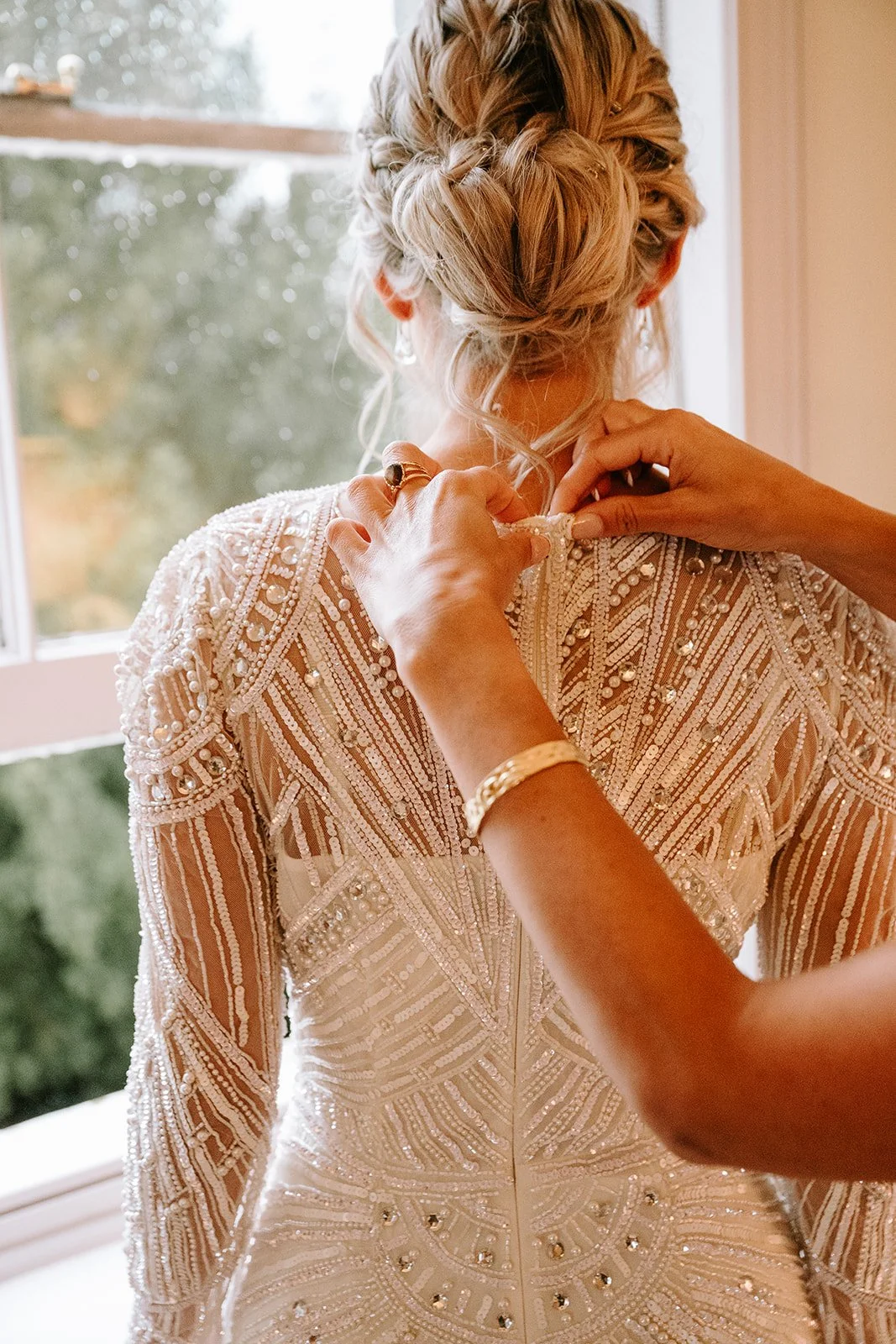 A bride is getting dressed, with someone helping her button up an intricately beaded and sequined wedding dress, standing near a window with blurred greenery outside.