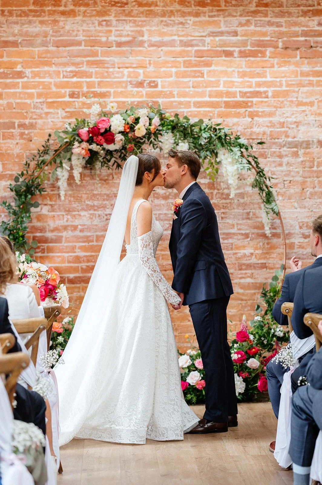 A bride and groom kiss during their wedding ceremony, standing in front of a floral arch against a brick wall, with guests seated on either side.