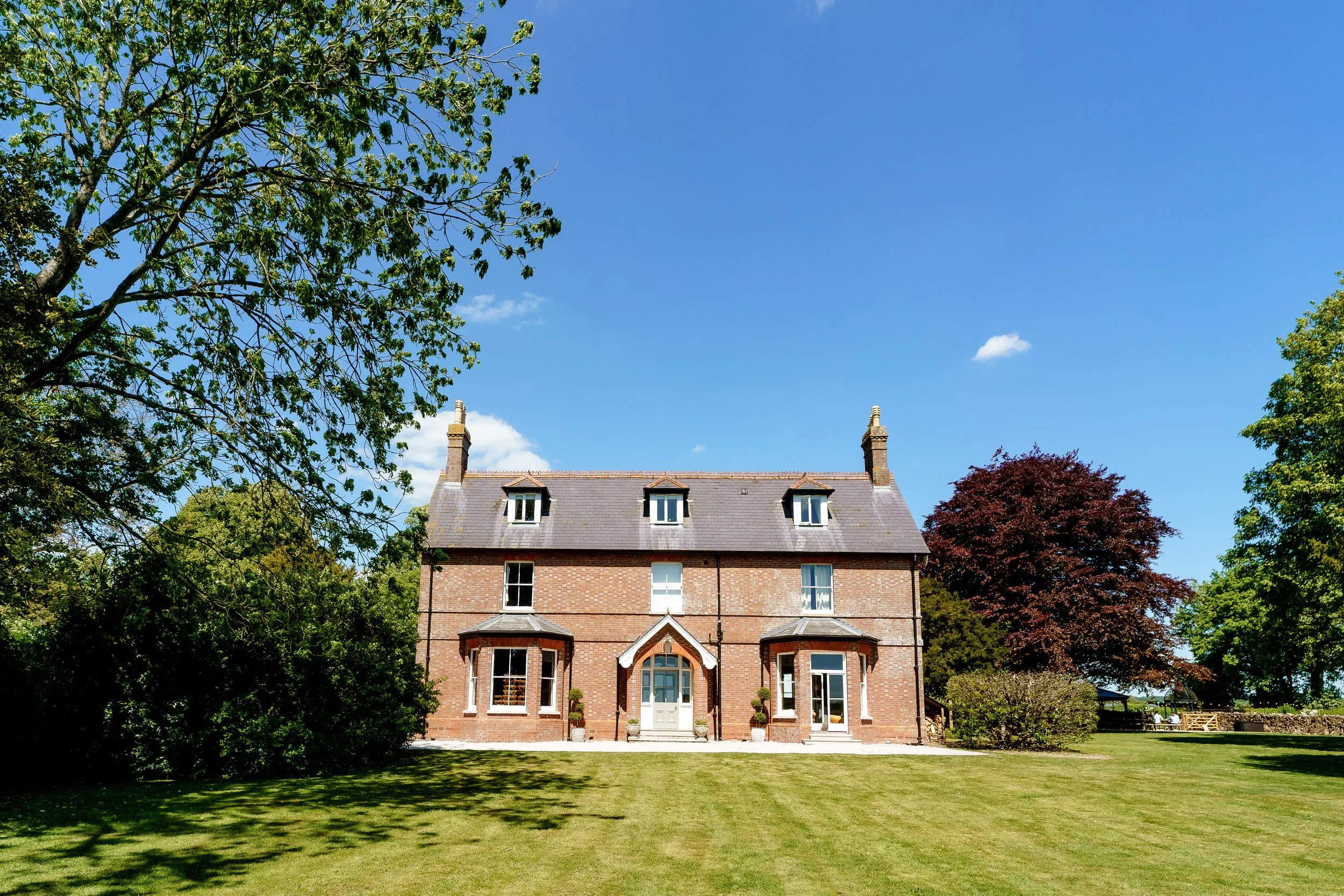 A brick house with a gray roof, multiple windows, and a white front door, surrounded by green trees and a well-kept lawn under a bright blue sky.