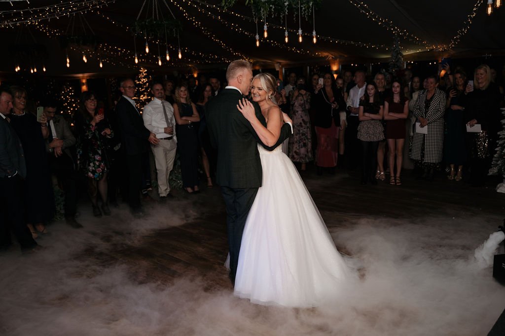 Bride and groom dancing at a wedding reception, surrounded by guests under string lights.
