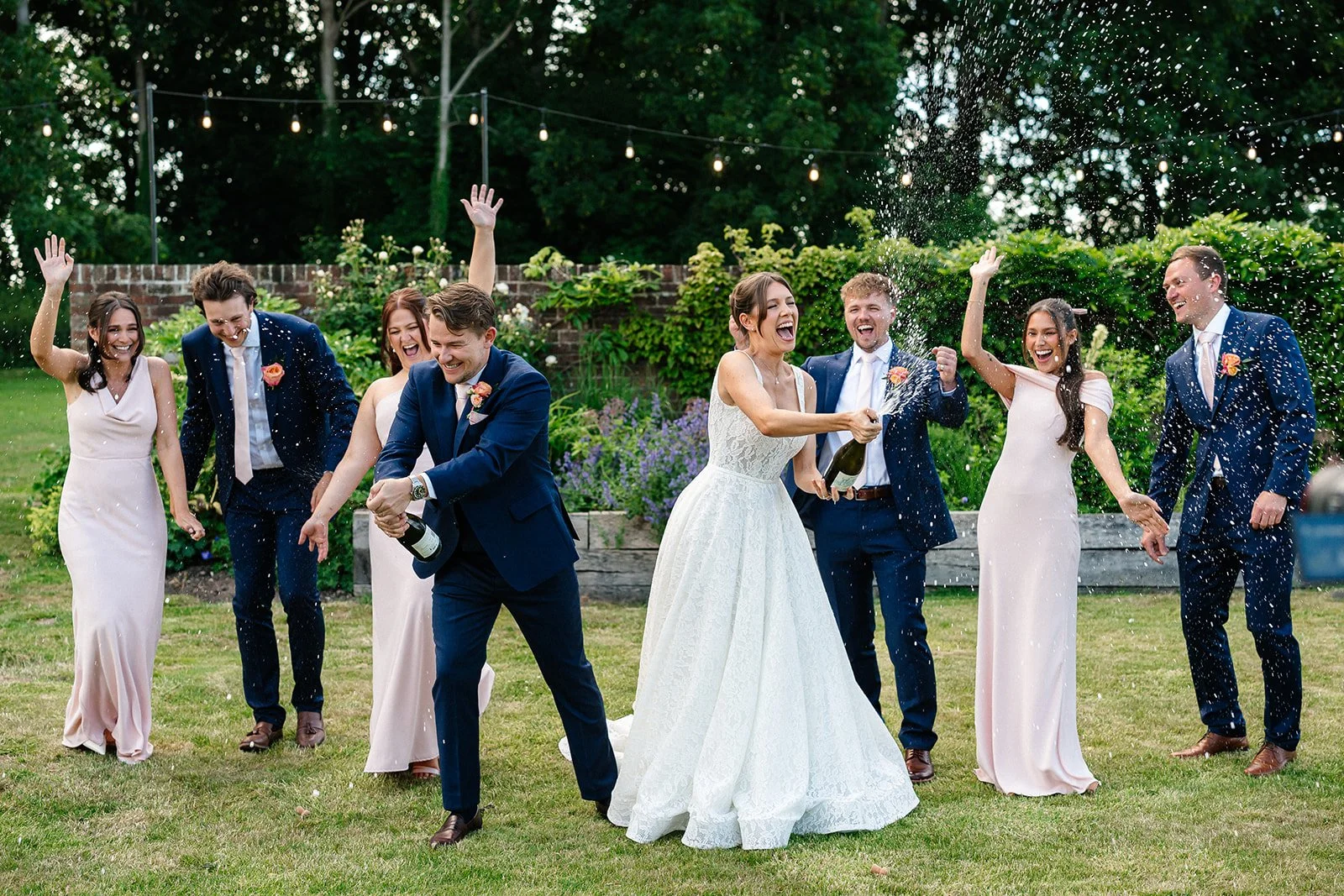 People celebrating a wedding outdoors, with the bride and groom in the center, opening a champagne bottle, and friends around them smiling and spraying champagne.
