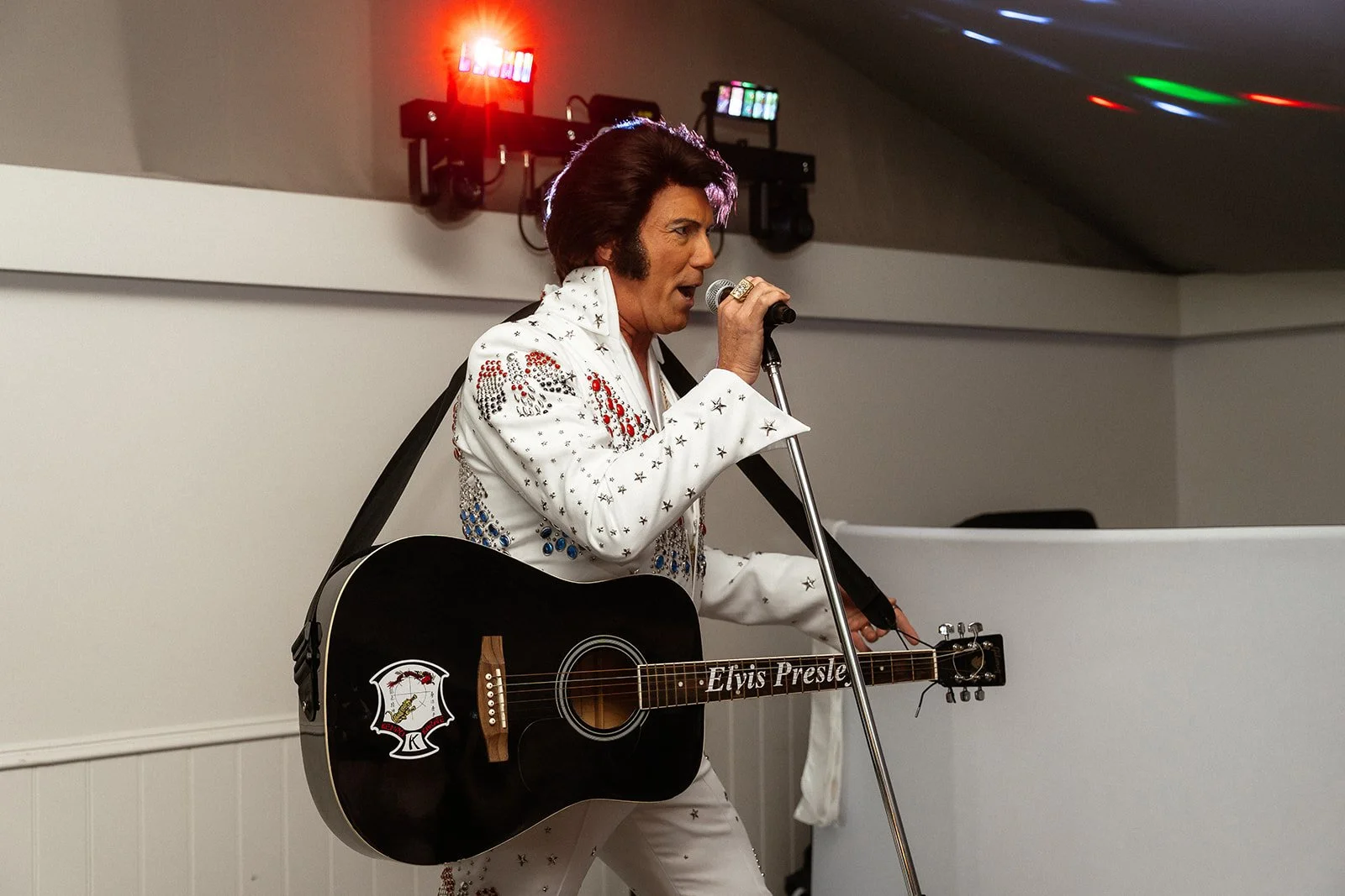 A person dressed as Elvis Presley performing with a black guitar, wearing a white jumpsuit decorated with red, blue, and black stars and diamonds, singing into a vintage microphone.