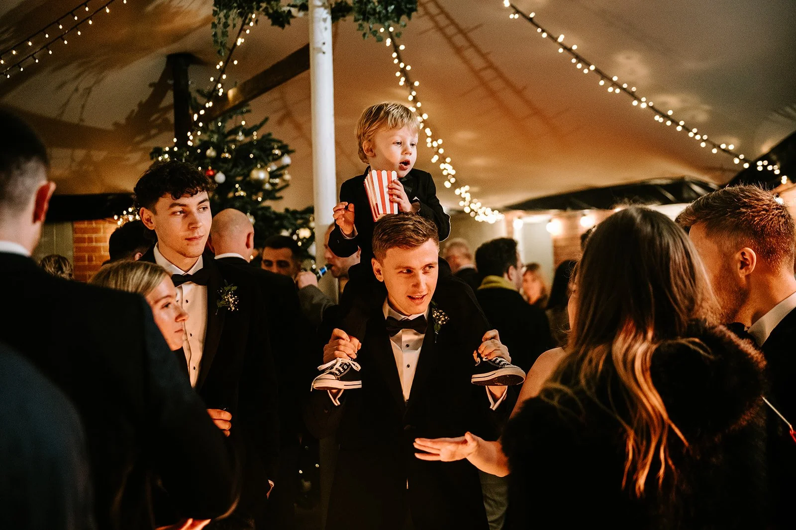 People in formal attire at a celebration, with a Christmas tree and festive string lights in the background, and a young boy sitting on an adult's shoulders holding popcorn.