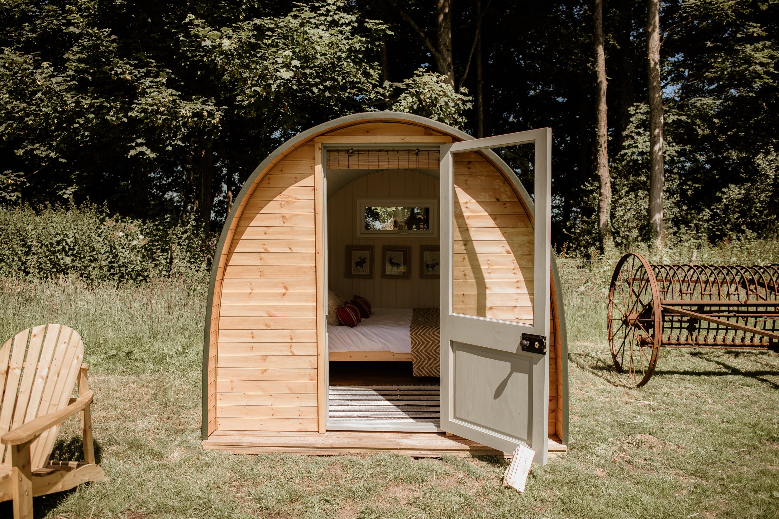 Small wooden cabin with open door in a grassy field, surrounded by trees and vintage farming equipment.