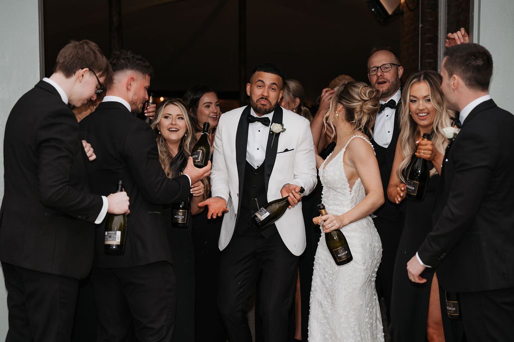 Wedding celebration with bride, groom, and guests in formal attire holding champagne bottles.