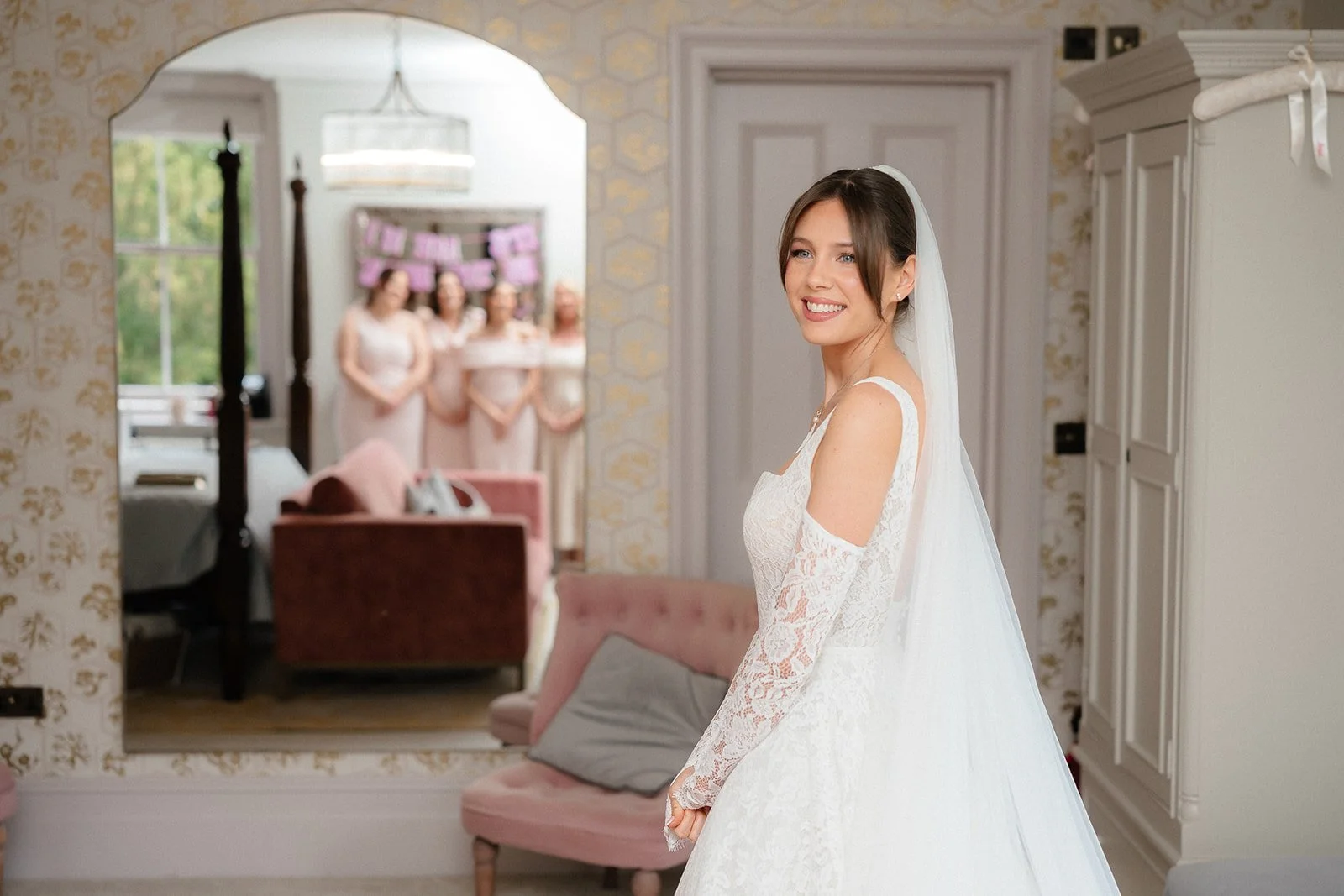 A bride in a white lace wedding dress smiling in a room, with her bridesmaids visible in the background through a mirror.