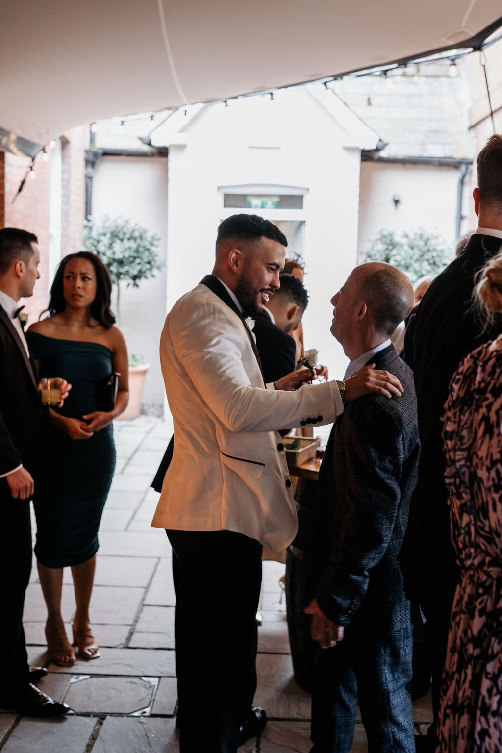 People in formal attire socializing at an event, featuring a man in a white suit jacket smiling and interacting with others.