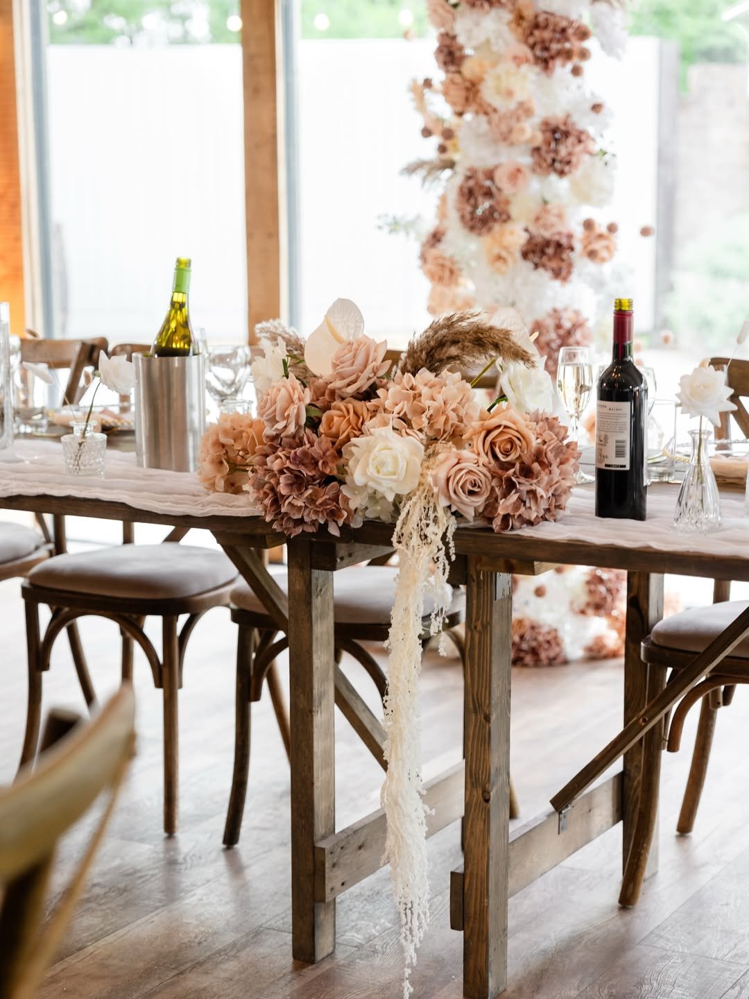 A wooden dining table decorated with a large floral centerpiece of blush pink, cream, and light brown roses and hydrangeas, and some dried flowers. There are wine bottles, glasses, and small vases on the table. In the background, there is an outdoor 