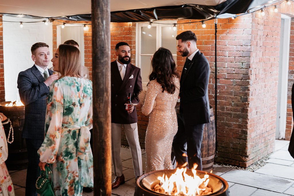 Group of people in formal attire socializing near a fire pit, with brick walls and string lights in the background.