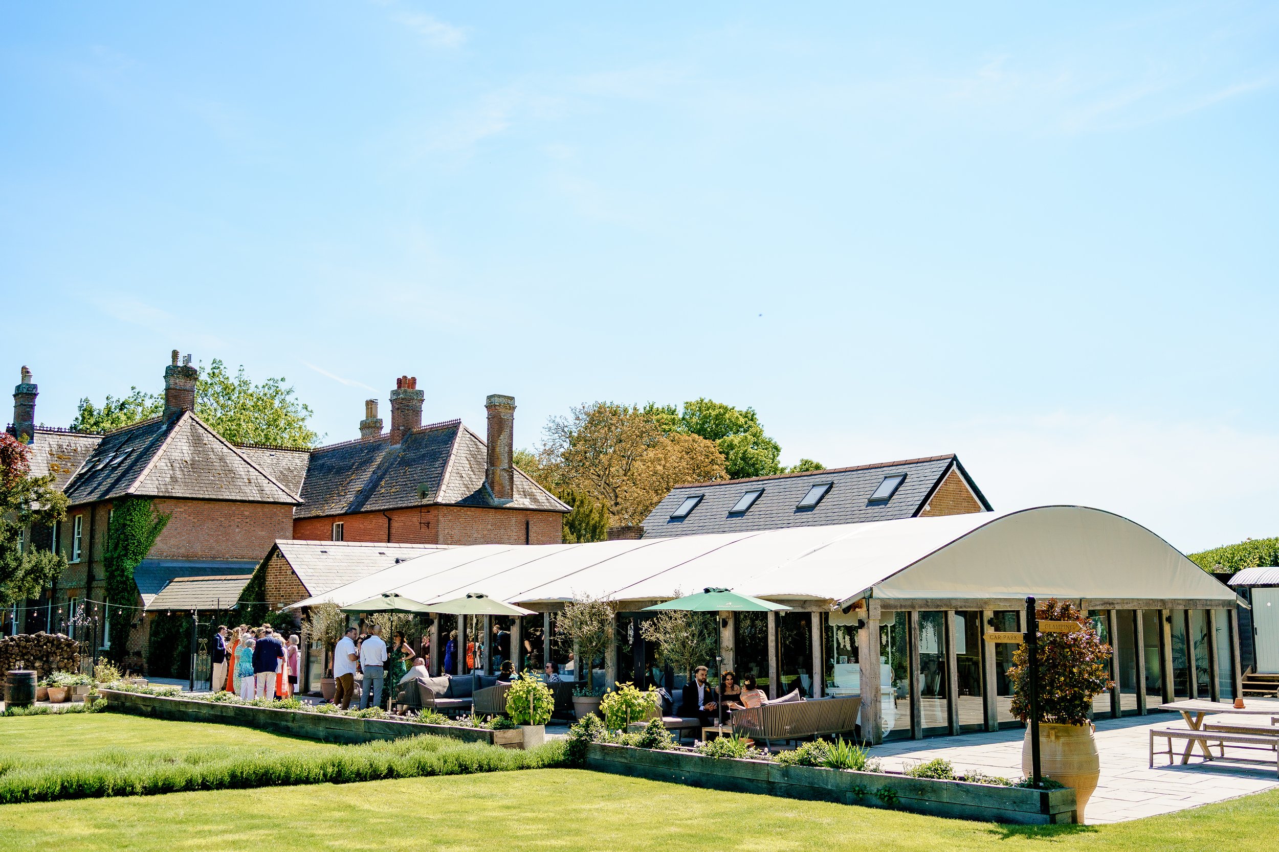 Outdoor gathering at a modern event space with a large white canopy, surrounded by a well-manicured lawn and neighboring traditional brick houses under a clear blue sky.