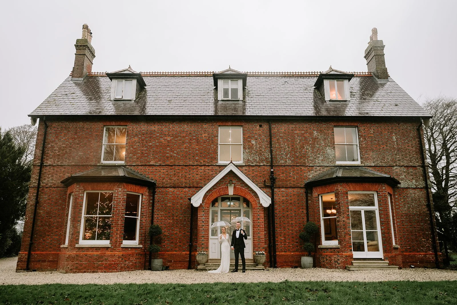 A bride and groom standing outside a large brick house holding clear umbrellas on a rainy day.
