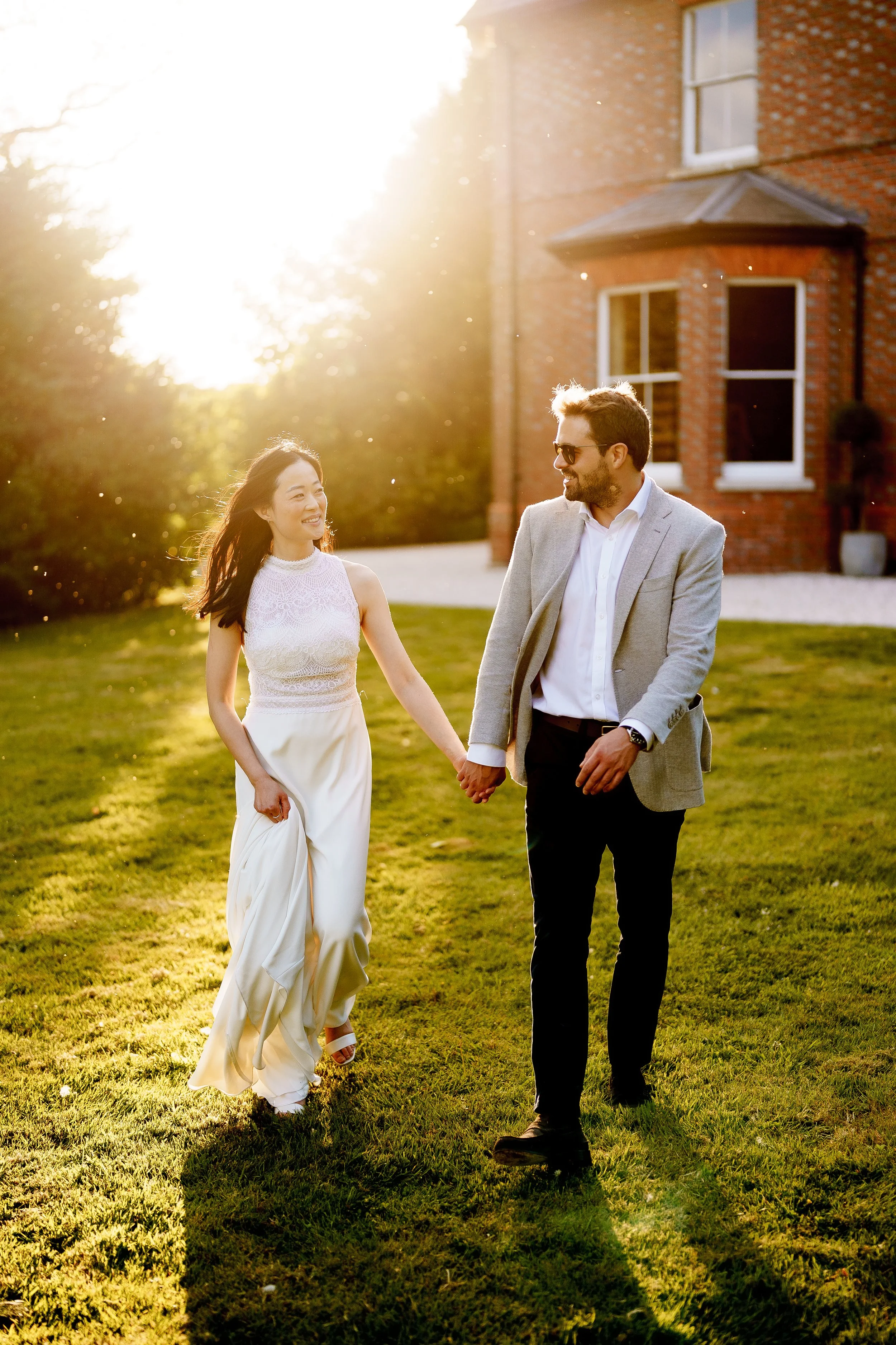 A couple, a woman in a white dress and a man in a grey blazer, holding hands and walking on a grassy lawn in front of a brick house during sunset.