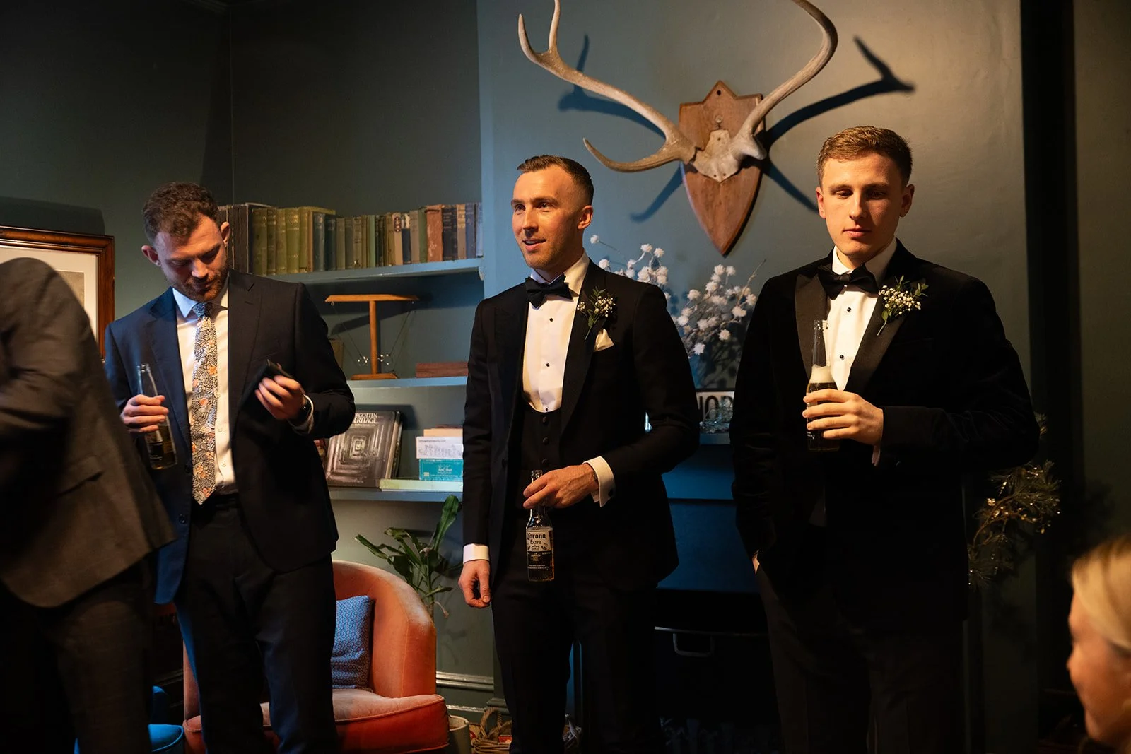 Three men in tuxedos at a formal gathering, standing with drinks, one in the middle speaking, all in a room with green walls, books, a deer head mounted on the wall, and decorative plants.
