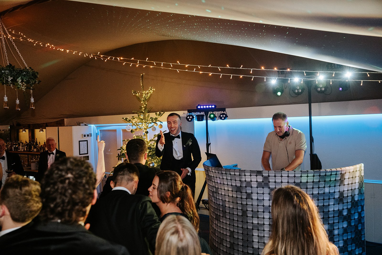 A group of people at a party, with a man in a tuxedo smiling and giving a thumbs-up, a DJ with headphones, and a decorated Christmas tree in the background, under string lights inside a tent.