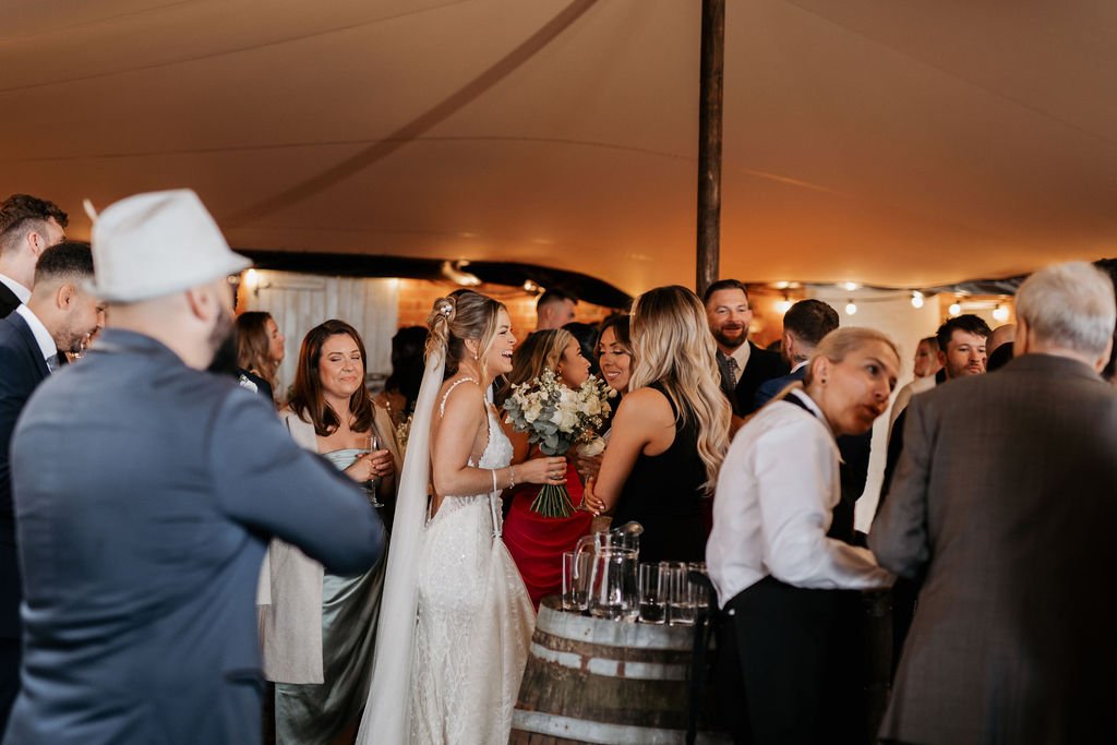 Guests mingling at a wedding reception under a tent, with a bride in a white dress holding flowers, surrounded by friends and a bartender in the foreground.