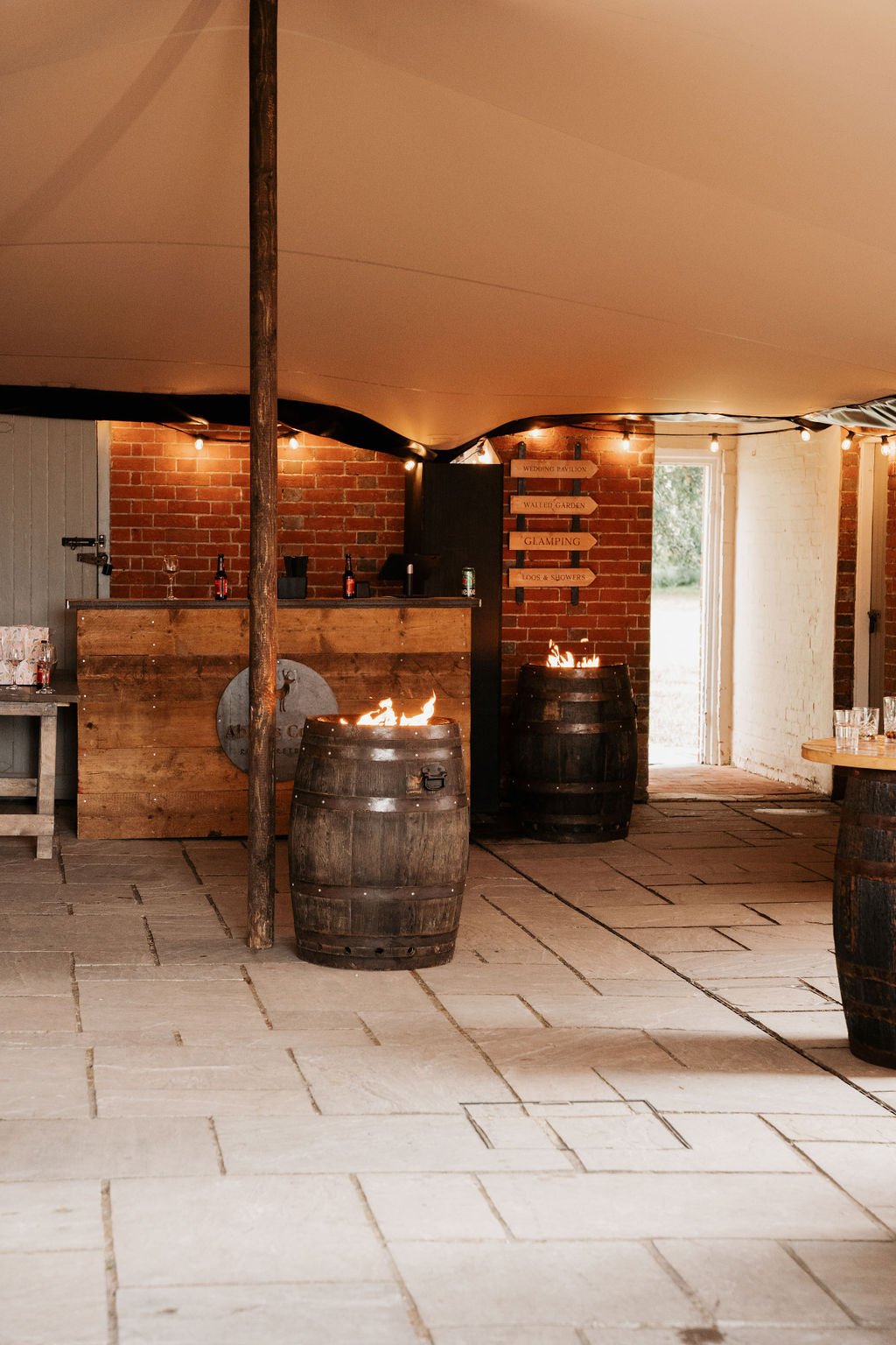 Rustic indoor bar setup with brick walls, wooden bar counter, and barrel fire pits; signs point to various areas like 'Wedding Pavilion' and 'Glamping.'