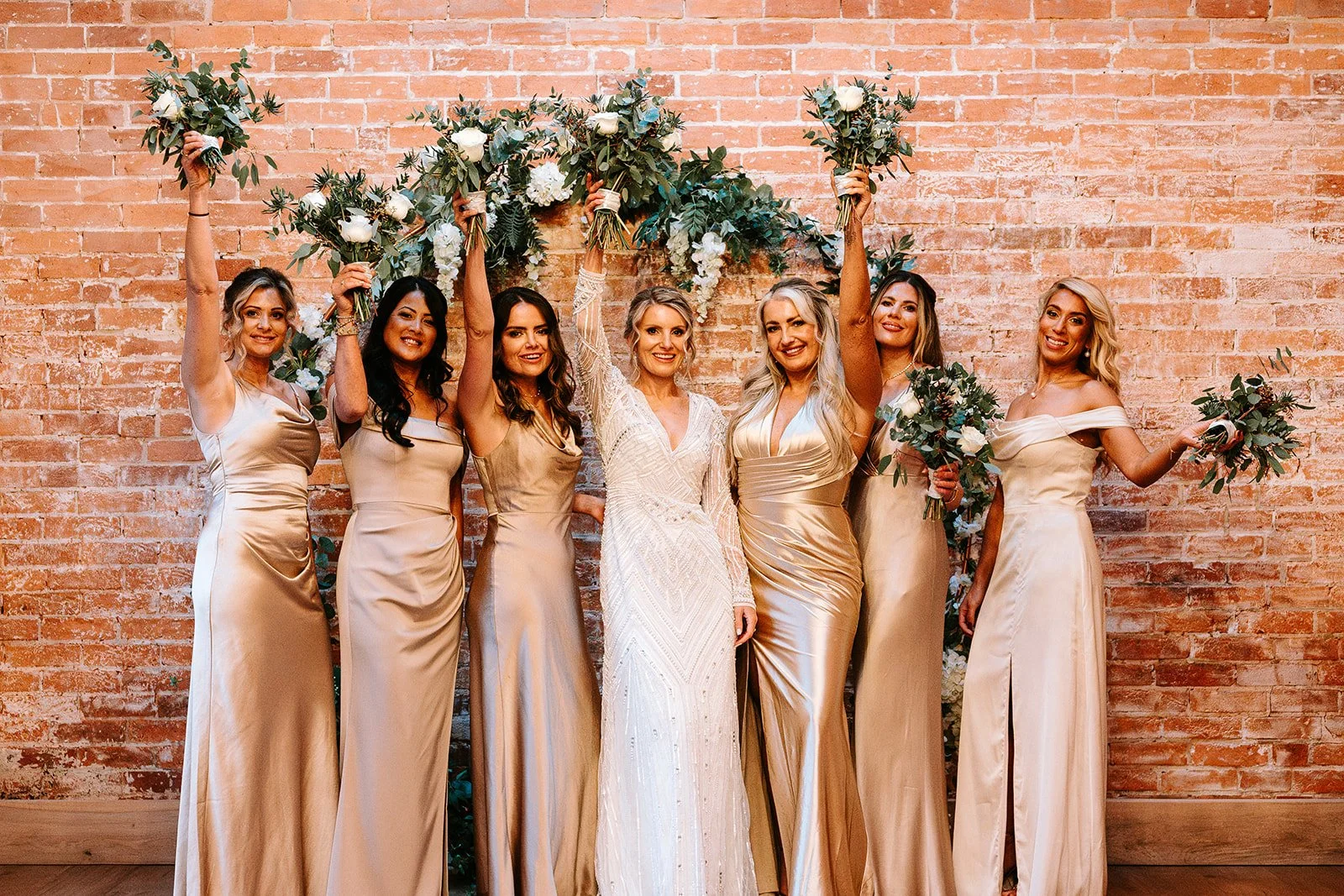 A group of seven women in elegant dresses standing in front of a brick wall with floral decorations, celebrating a wedding. Some of the women are holding bouquets and raising their hands.