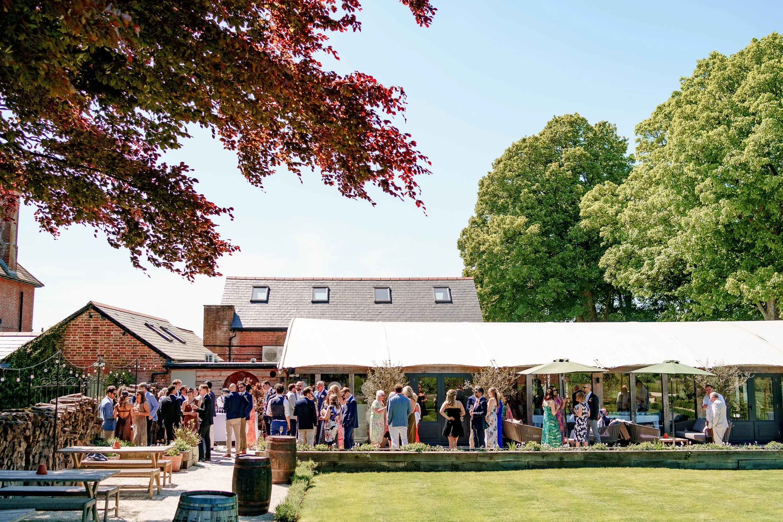 People gathered outdoors at a social event with a large white tent, surrounded by green trees and a lawn, on a sunny day.