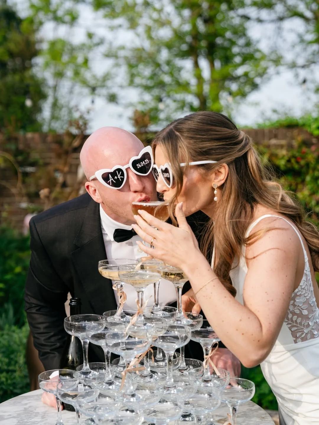 A couple at a wedding kissing over a tower of champagne glasses, both wearing large heart-shaped sunglasses with wedding details and initials.