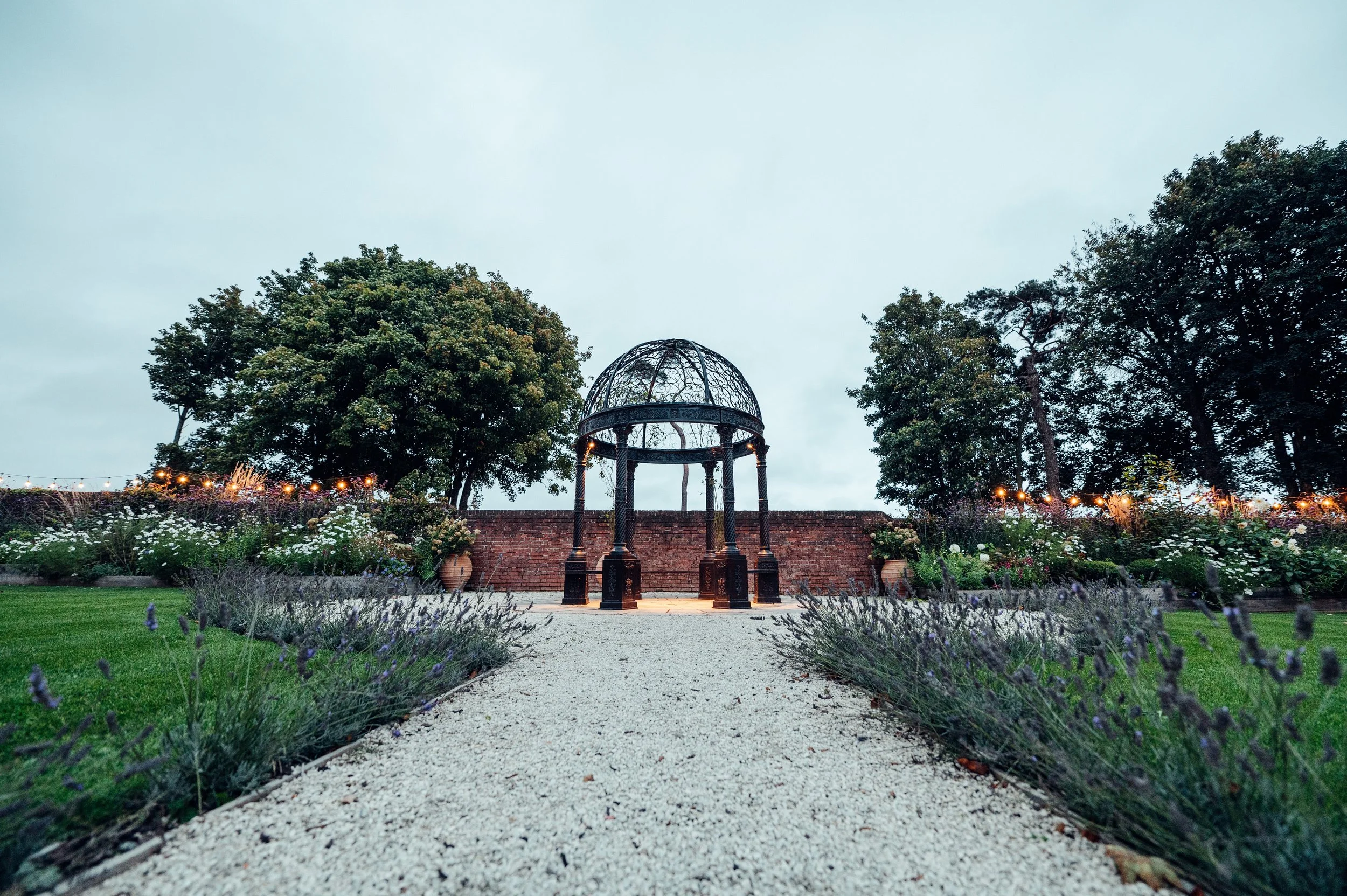 Garden landscape with metal gazebo, stone pathway, lush greenery and flowers, string lights, and trees in the background.