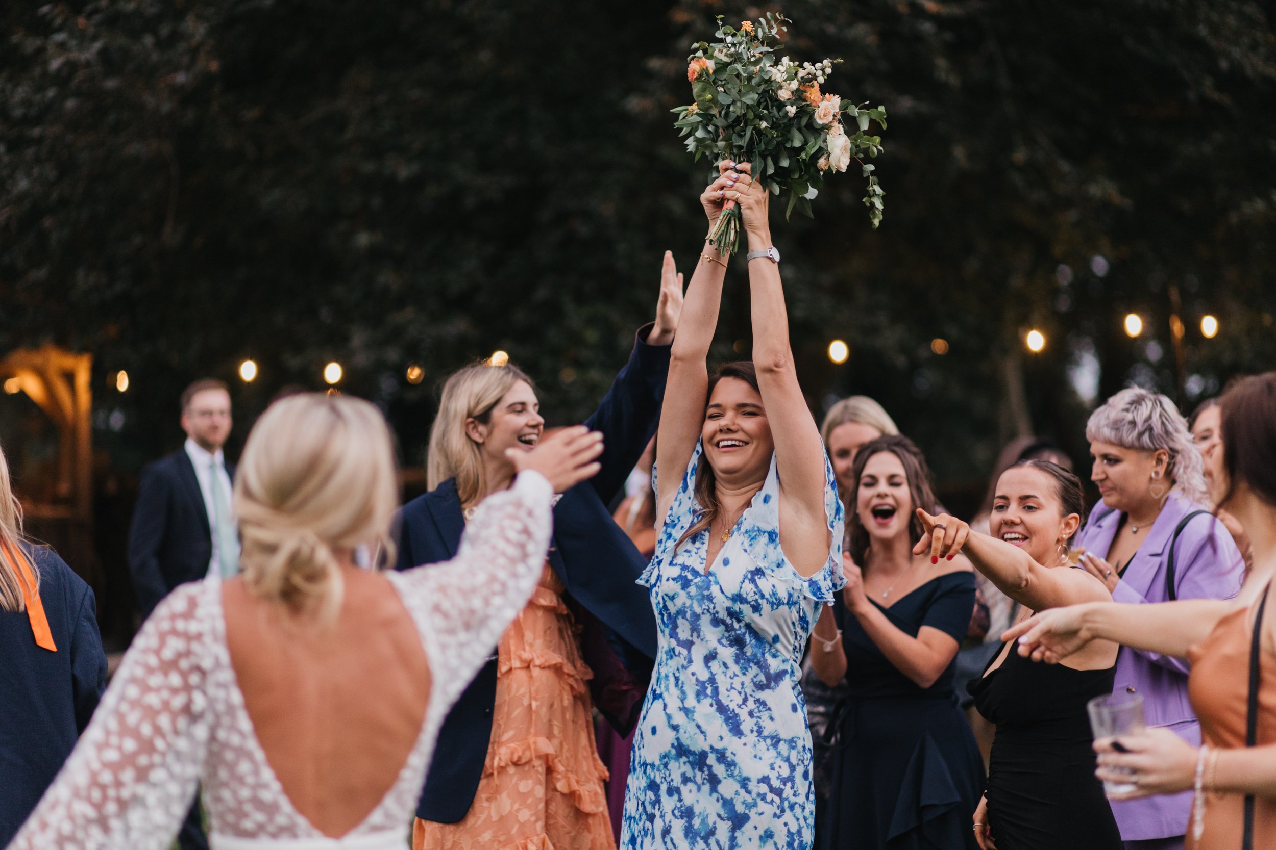 Group of women celebrating at a wedding, one holding a bouquet, outdoors.