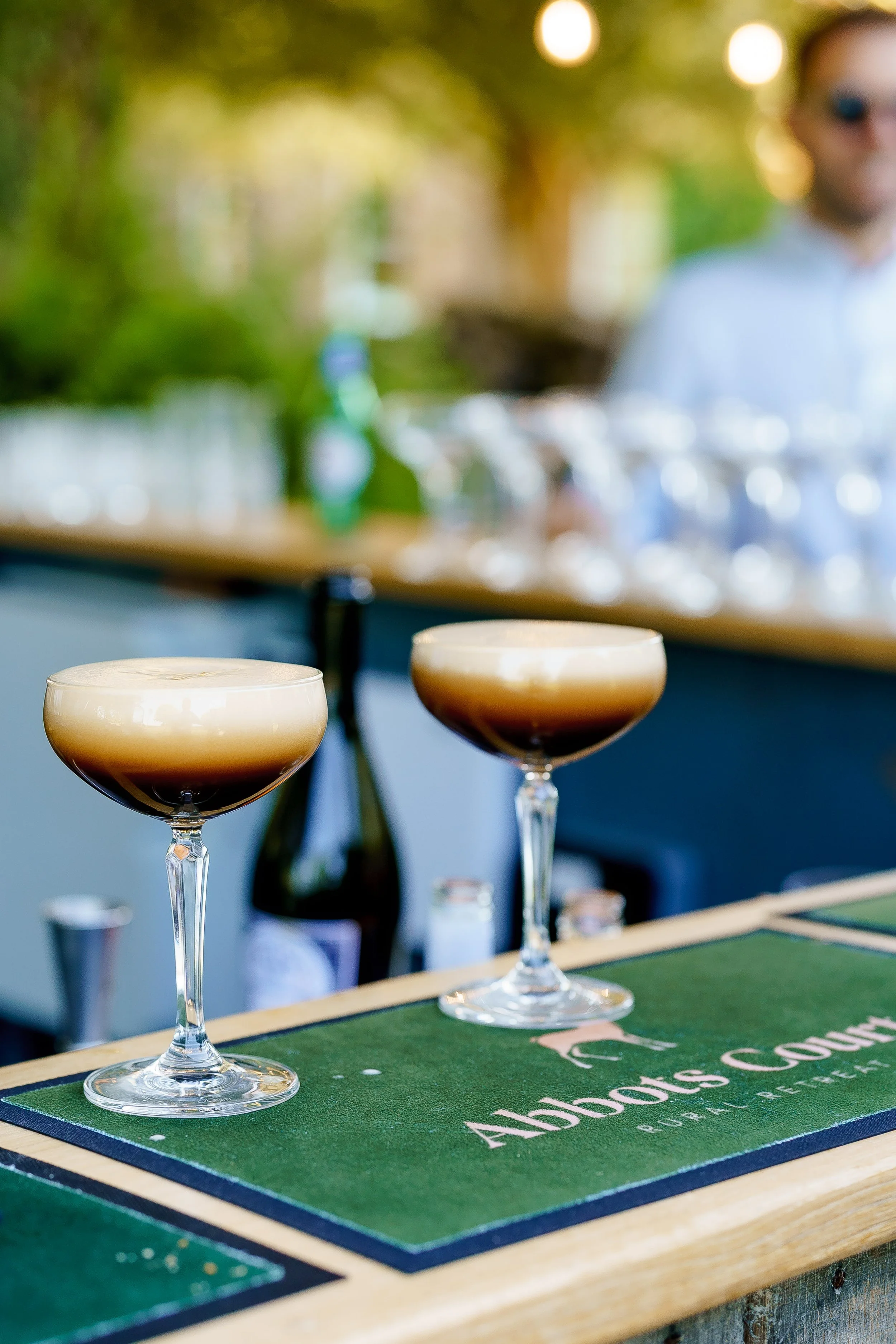 Two cocktails with layered foam and dark liquid in coupe glasses on a green bar mat with 'Abbots County' printed on it, in an outdoor setting.
