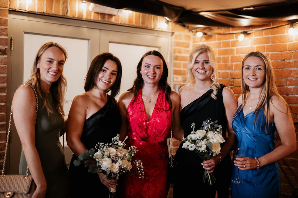 Five women in formal dresses posing indoors with brick wall in the background.
